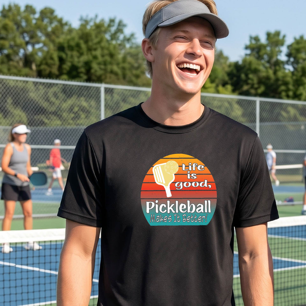 A smiling man wearing a black t-shirt with a retro pickleball graphic and the text 'Life is good, Pickleball Makes It Better' on an outdoor pickleball court.