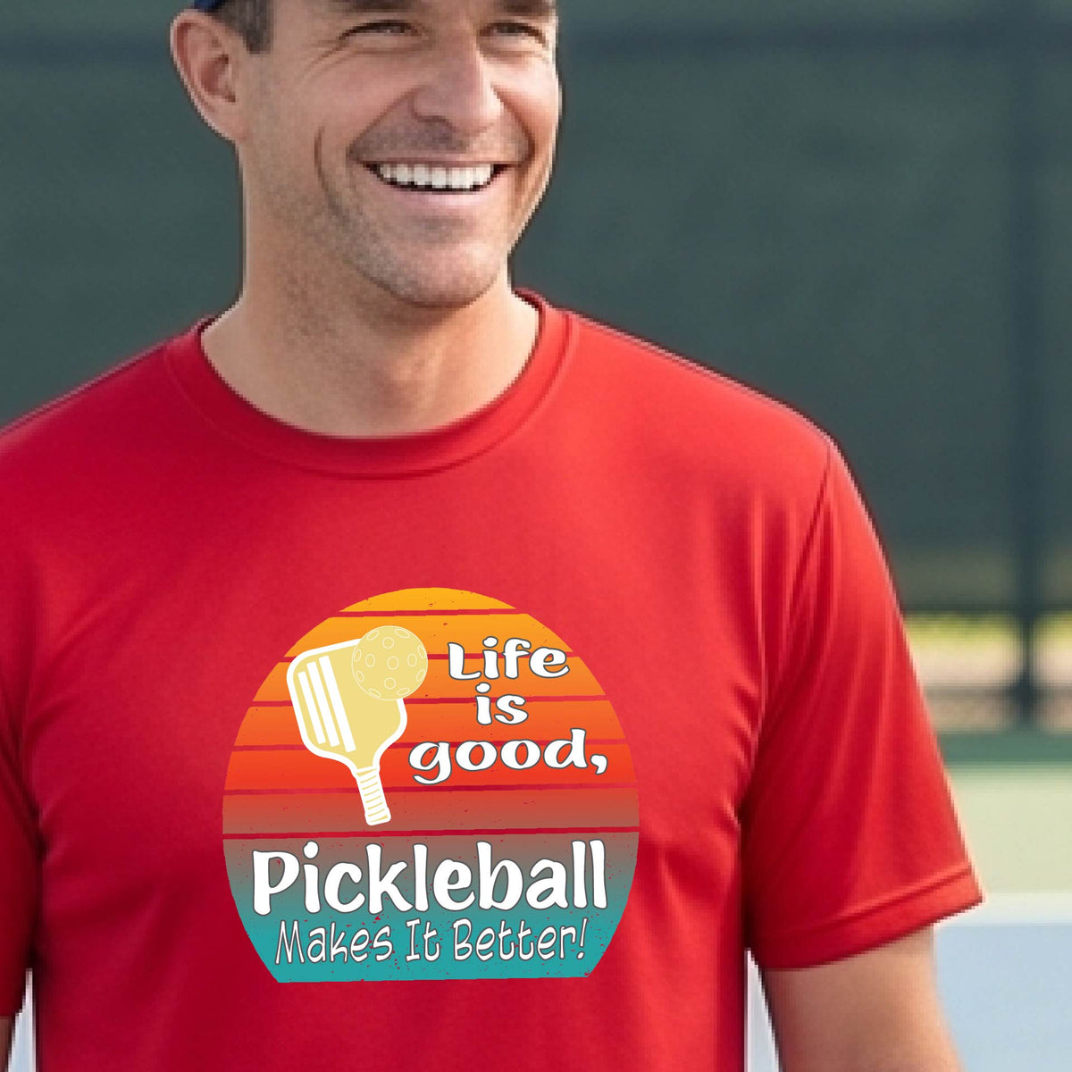 A smiling man wearing a red t-shirt with a retro pickleball graphic and the text 'Life is good, Pickleball Makes It Better' on an outdoor pickleball court.