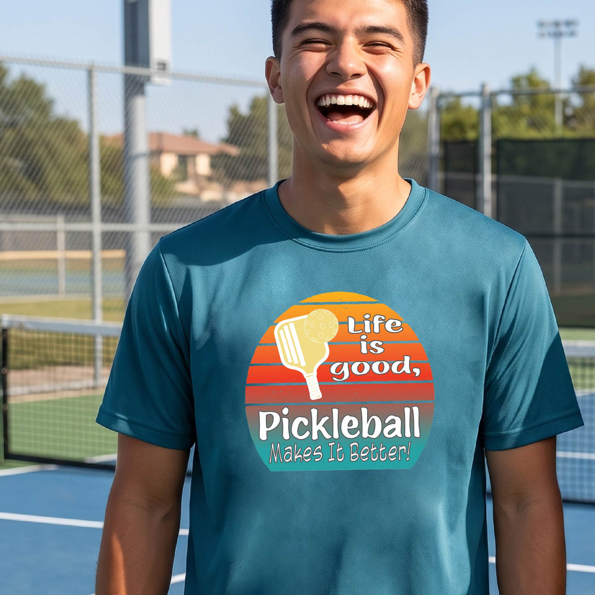 A smiling man wearing a teal blue t-shirt with a retro pickleball graphic and the text 'Life is good, Pickleball Makes It Better' on an outdoor pickleball court.