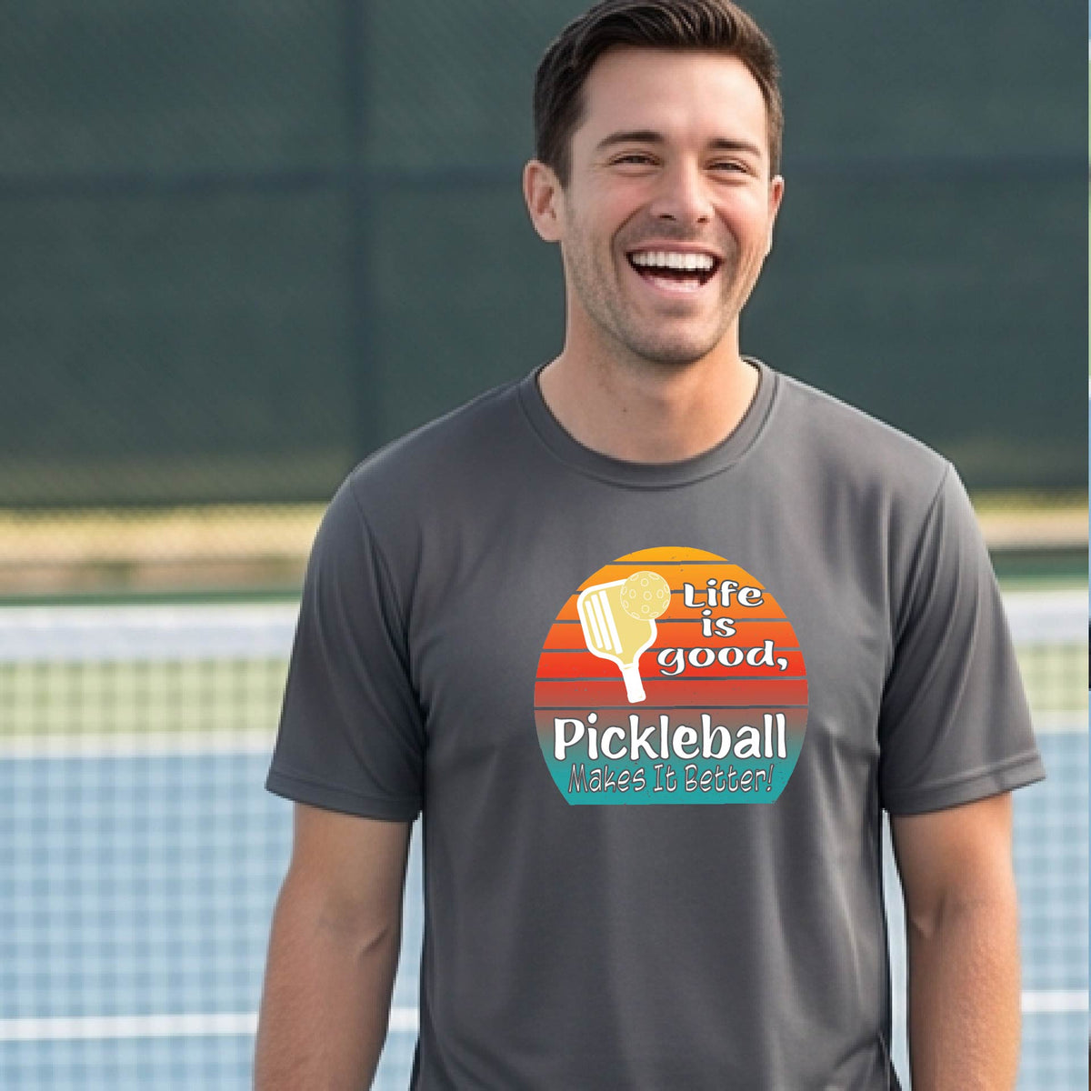 A smiling man wearing a dark gray t-shirt with a retro pickleball graphic and the text 'Life is good, Pickleball Makes It Better' on an outdoor pickleball court.