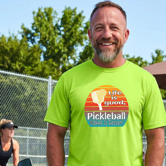 A smiling man wearing a bright lime green t-shirt with a retro pickleball graphic and the text 'Life is good, Pickleball Makes It Better' on an outdoor pickleball court.