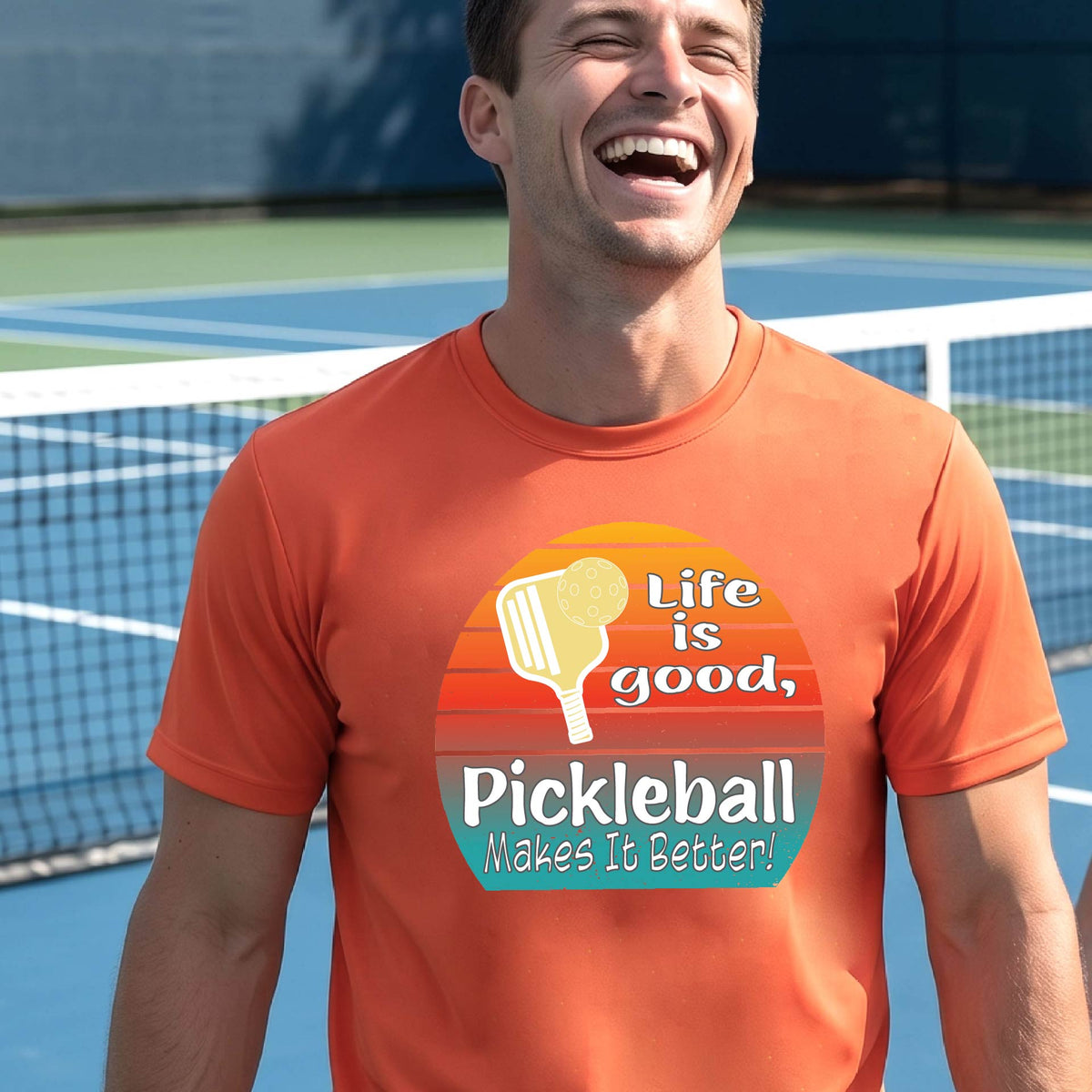A smiling man wearing a bright orange t-shirt with a retro pickleball graphic and the text 'Life is good, Pickleball Makes It Better' on an outdoor pickleball court.