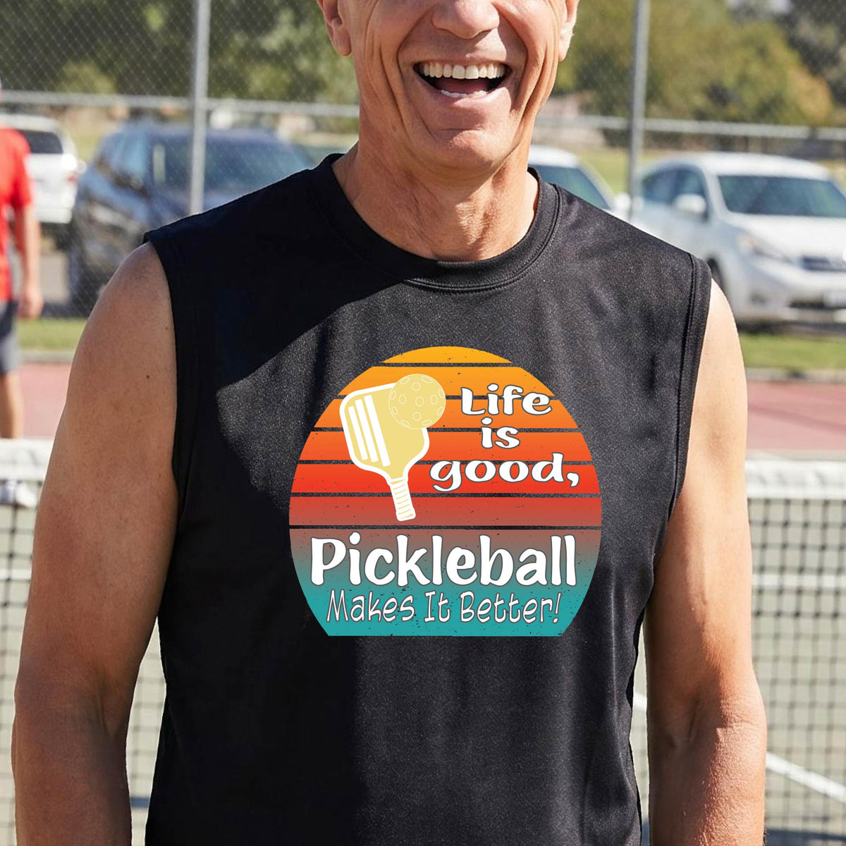 A man wearing a black sleeveless t-shirt with a retro multi-colored circle with a pickleball paddle and ball, with white text that says 'LIFE IS GOOD PICKLEBALL MAKES IT BETTER', standing in front of a pickleball court.