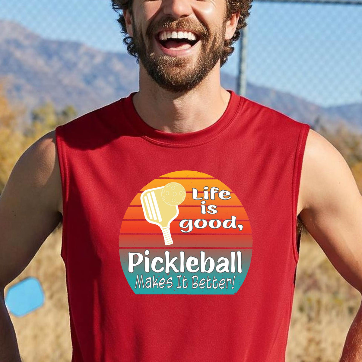 A man wearing a red sleeveless t-shirt with a retro multi-colored circle with a pickleball paddle and ball, with white text that says 'LIFE IS GOOD PICKLEBALL MAKES IT BETTER', standing in front of a pickleball court.