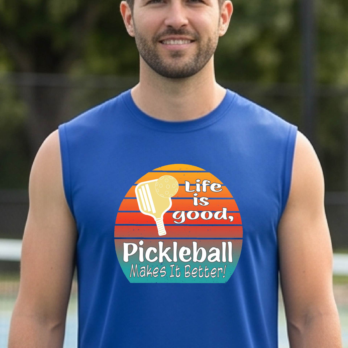 A man wearing a royal blue sleeveless t-shirt with a retro multi-colored circle with a pickleball paddle and ball, with white text that says 'LIFE IS GOOD PICKLEBALL MAKES IT BETTER', standing in front of a pickleball court.