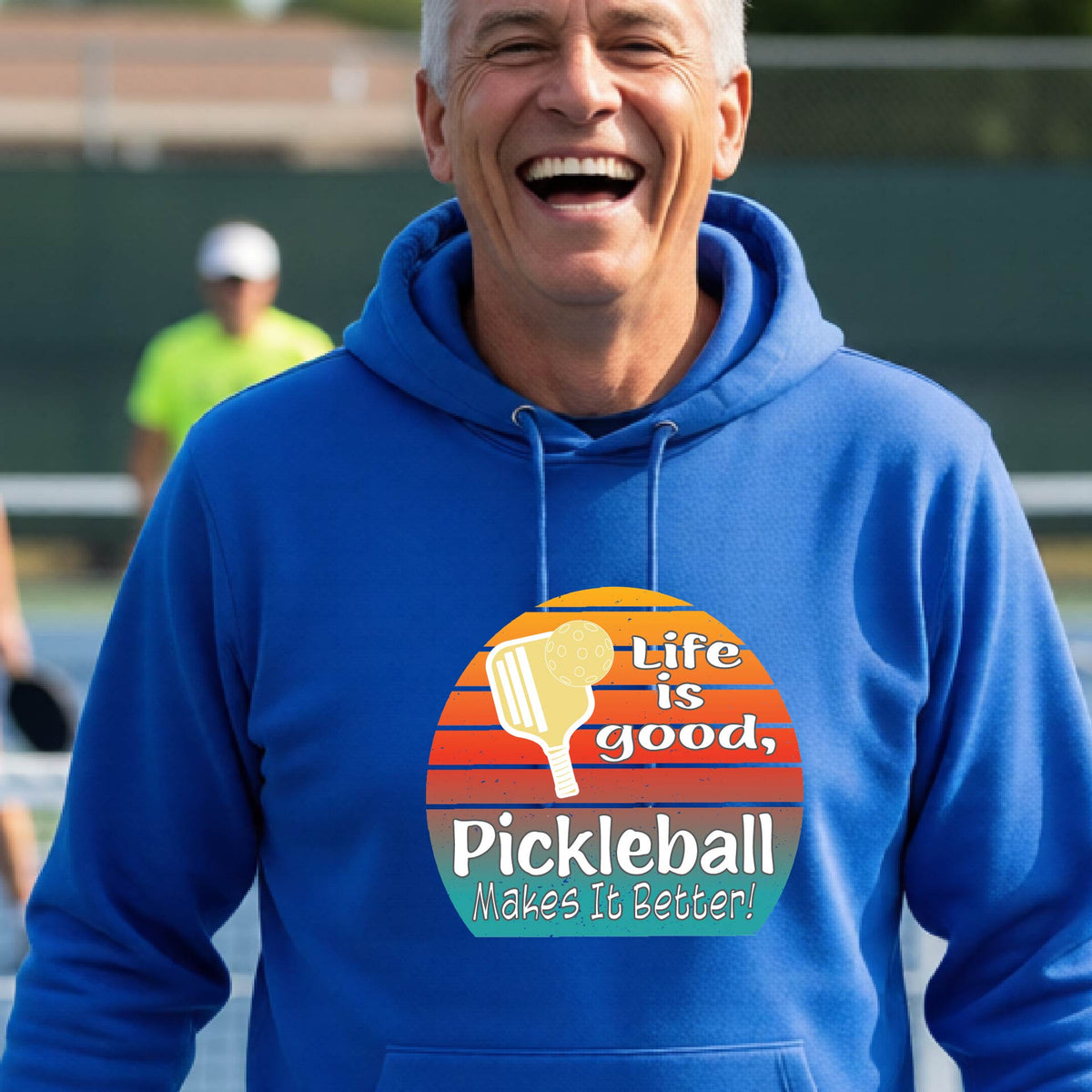 A man wearing a royal blue hoodie with a retro-style pickleball graphic on the chest, with the text 'LIFE IS GOOD PICKLEBALL MAKES IT BETTER' standing on an outdoor pickleball court with a green fence in the background.