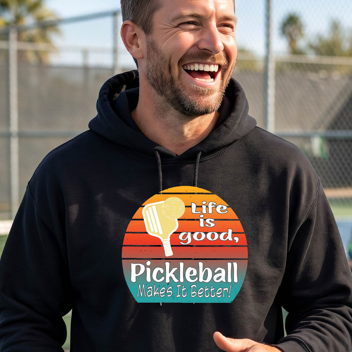 A man wearing a black hoodie with a retro-style pickleball graphic on the chest, with the text 'LIFE IS GOOD PICKLEBALL MAKES IT BETTER' standing on an outdoor pickleball court with a green fence in the background.