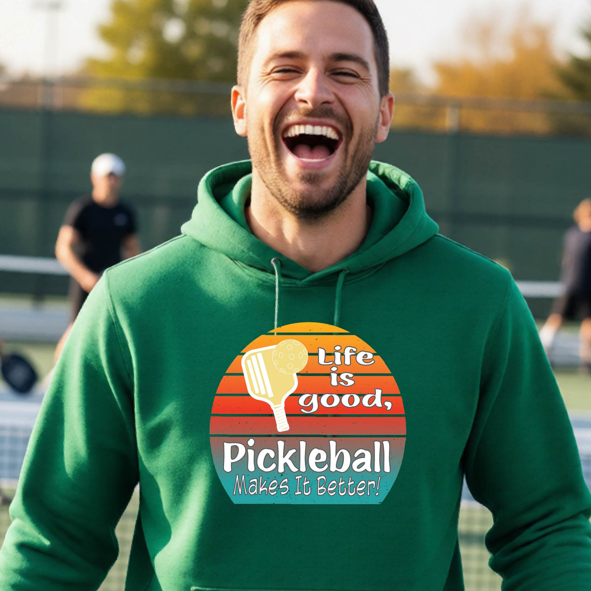 A man wearing a green hoodie with a retro-style pickleball graphic on the chest, with the text 'LIFE IS GOOD PICKLEBALL MAKES IT BETTER' standing on an outdoor pickleball court with a green fence in the background.