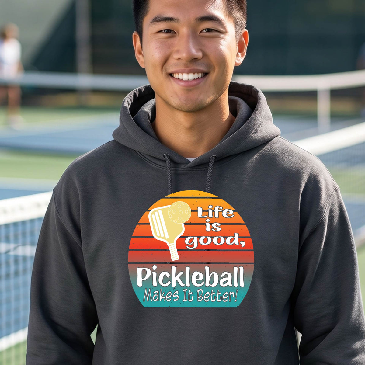 A man wearing a gray hoodie with a retro-style pickleball graphic on the chest, with the text 'LIFE IS GOOD PICKLEBALL MAKES IT BETTER' standing on an outdoor pickleball court with a green fence in the background.
