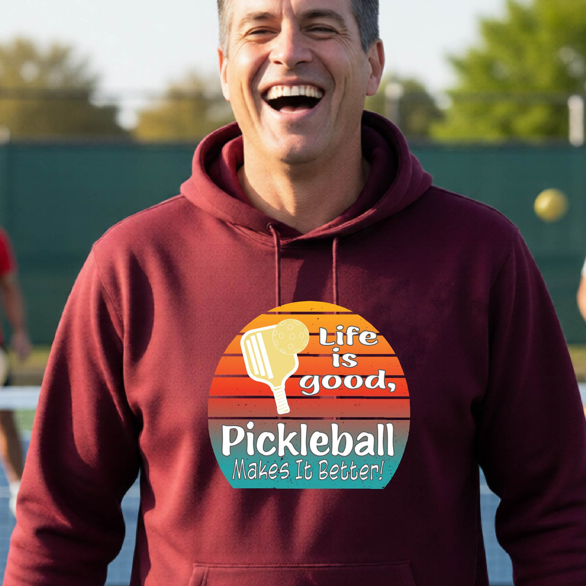 A man wearing a maroon hoodie with a retro-style pickleball graphic on the chest, with the text 'LIFE IS GOOD PICKLEBALL MAKES IT BETTER' standing on an outdoor pickleball court with a green fence in the background.