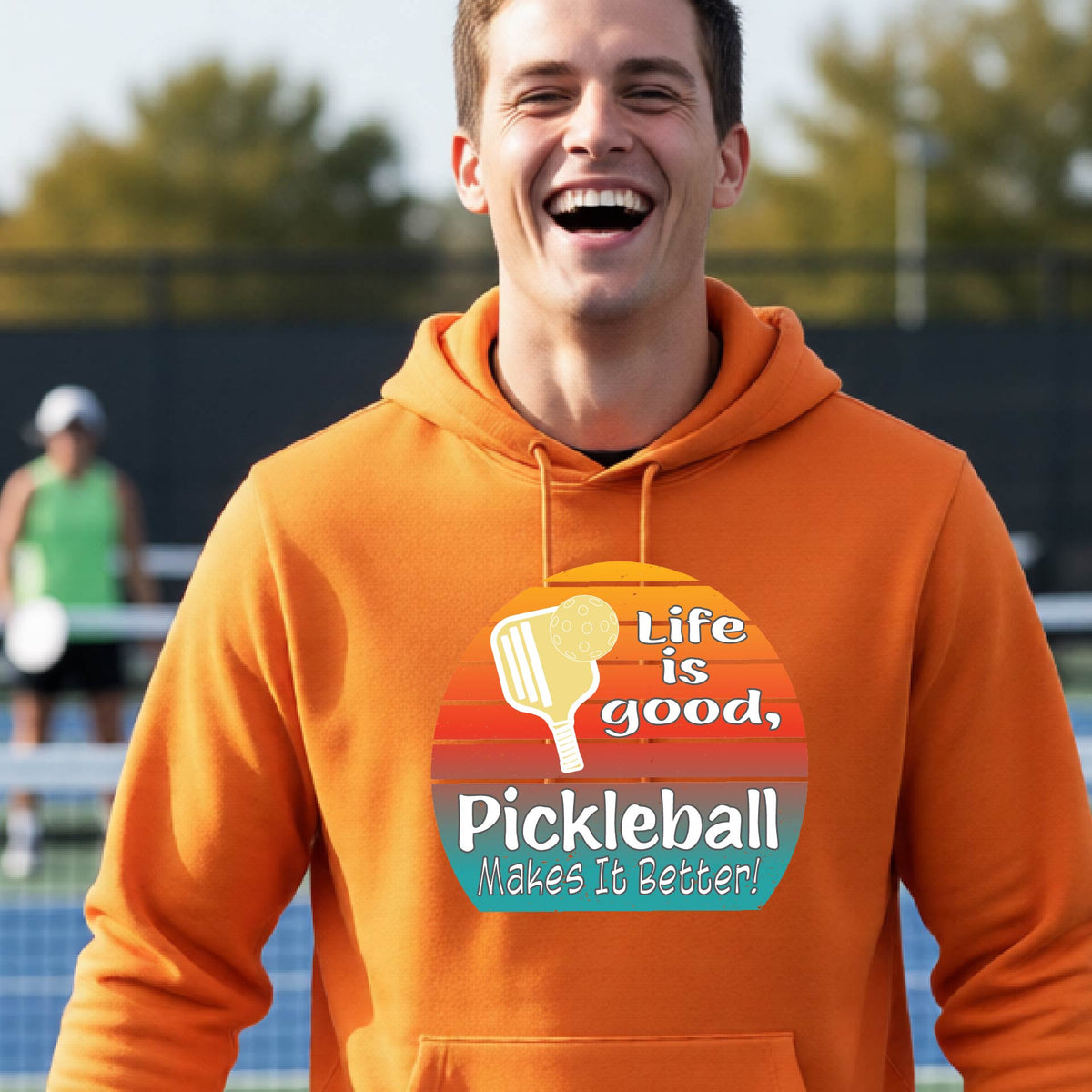 A man wearing an orange hoodie with a retro-style pickleball graphic on the chest, with the text 'LIFE IS GOOD PICKLEBALL MAKES IT BETTER' standing on an outdoor pickleball court with a green fence in the background.
