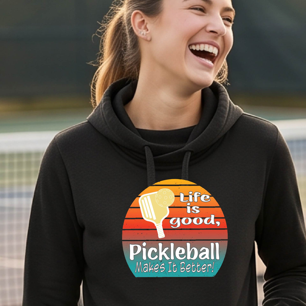 A woman wearing a black hoodie pullover sweatshirt with a retro multi-colored circle with a pickleball paddle and ball, with white text that says 'LIFE IS GOOD PICKLEBALL MAKES IT BETTER', standing in front of a pickleball court.