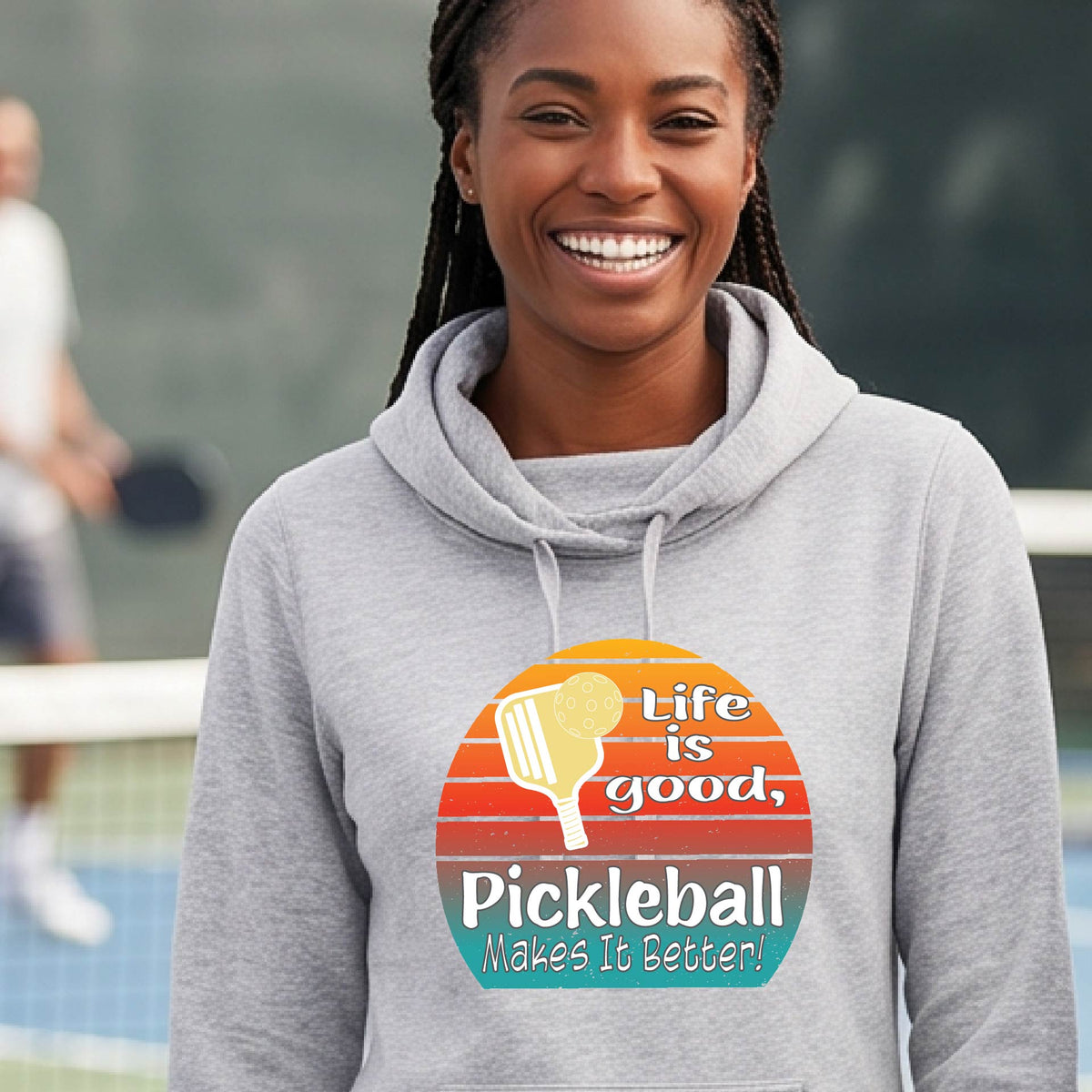 A woman wearing a light gray hoodie pullover sweatshirt with a retro multi-colored circle with a pickleball paddle and ball, with white text that says 'LIFE IS GOOD PICKLEBALL MAKES IT BETTER', standing in front of a pickleball court.