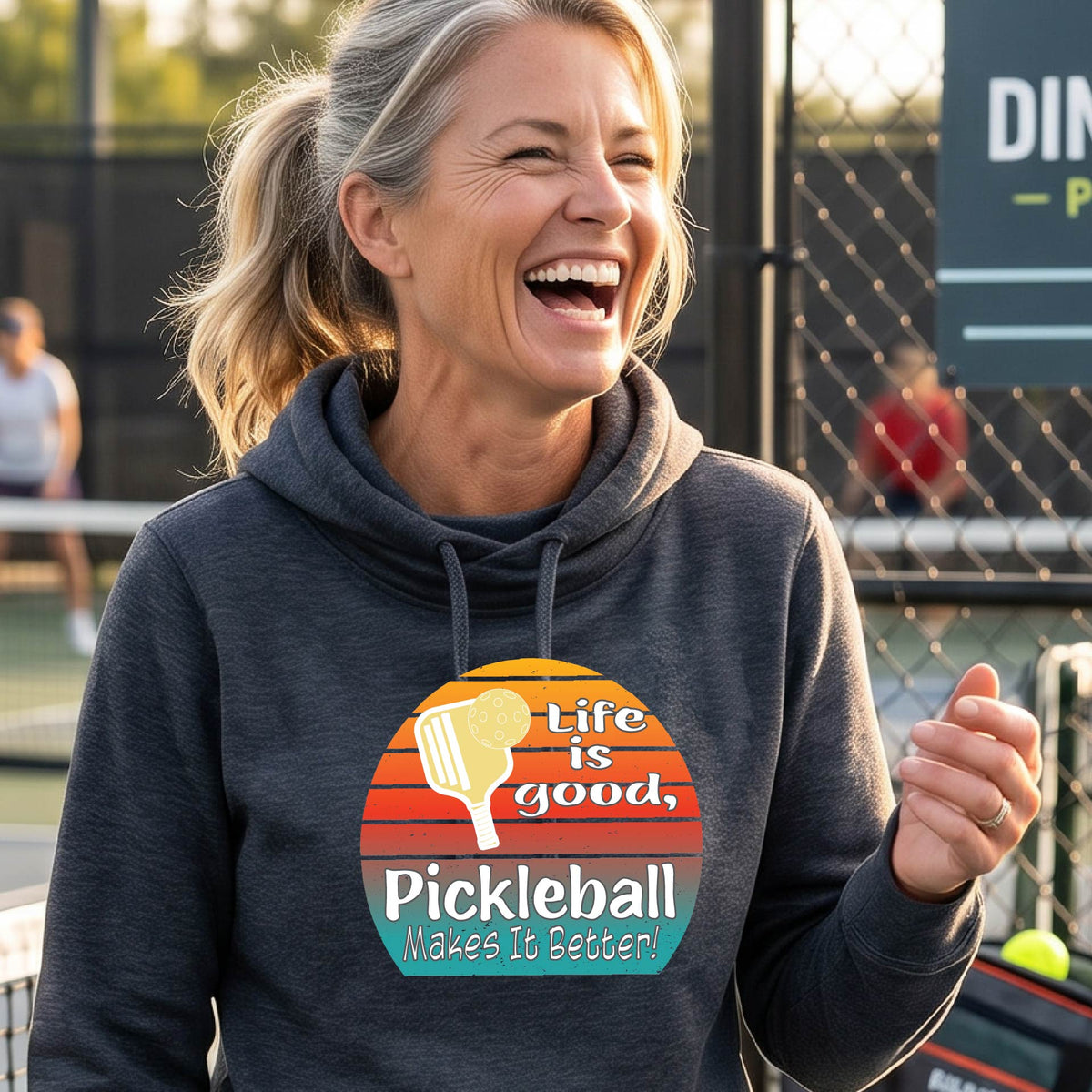 A woman wearing a navy blue hoodie pullover sweatshirt with a retro multi-colored circle with a pickleball paddle and ball, with white text that says 'LIFE IS GOOD PICKLEBALL MAKES IT BETTER', standing in front of a pickleball court.