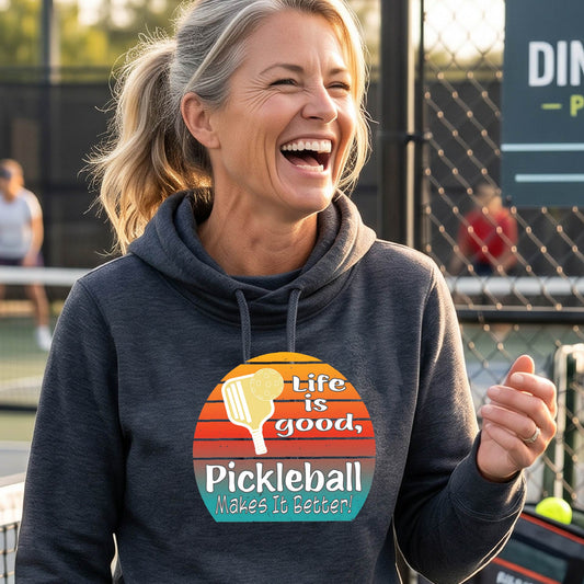 A woman wearing a navy blue hoodie pullover sweatshirt with a retro multi-colored circle with a pickleball paddle and ball, with white text that says 'LIFE IS GOOD PICKLEBALL MAKES IT BETTER', standing in front of a pickleball court.