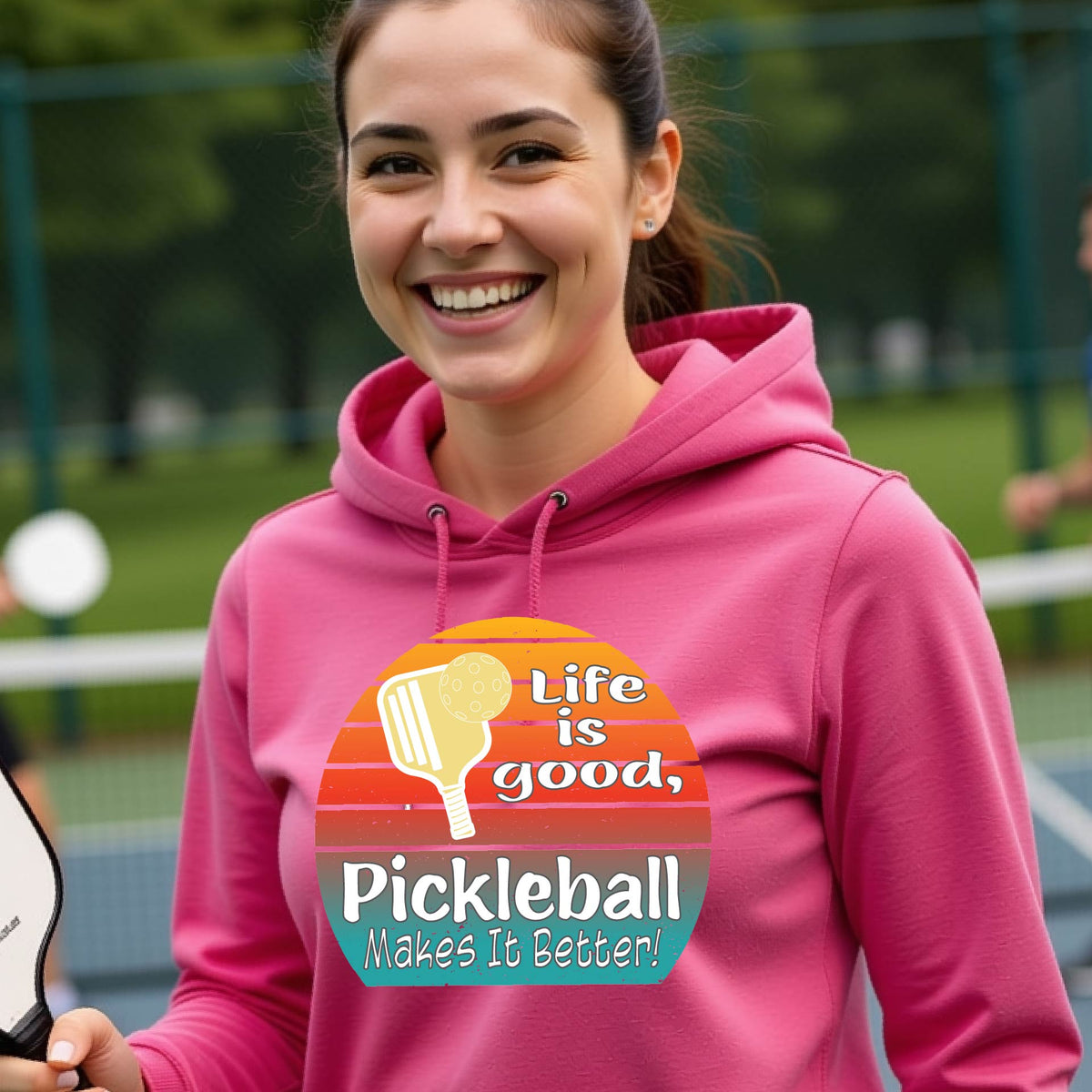 A woman wearing a pink hoodie pullover sweatshirt with a retro multi-colored circle with a pickleball paddle and ball, with white text that says 'LIFE IS GOOD PICKLEBALL MAKES IT BETTER', standing in front of a pickleball court.