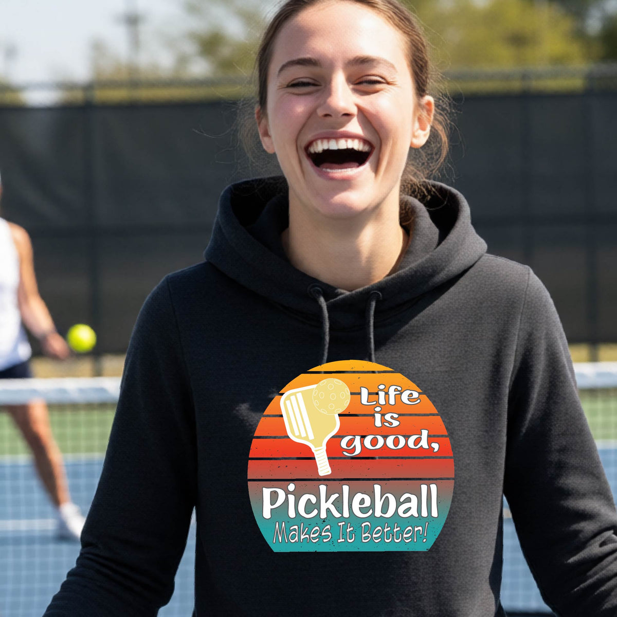 A woman wearing a black hoodie pullover with a retro multi-colored circle with a pickleball paddle and ball, with white text that says 'LIFE IS GOOD PICKLEBALL MAKES IT BETTER', standing in front of a pickleball court.