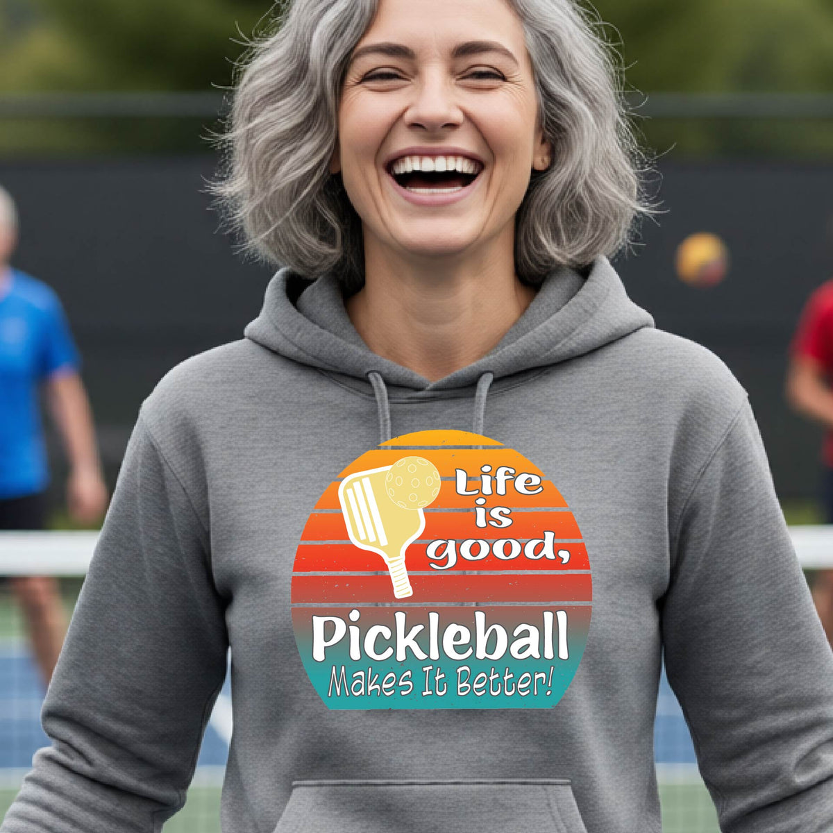 A woman wearing a dark gray hoodie pullover with a retro multi-colored circle with a pickleball paddle and ball, with white text that says 'LIFE IS GOOD PICKLEBALL MAKES IT BETTER', standing in front of a pickleball court.