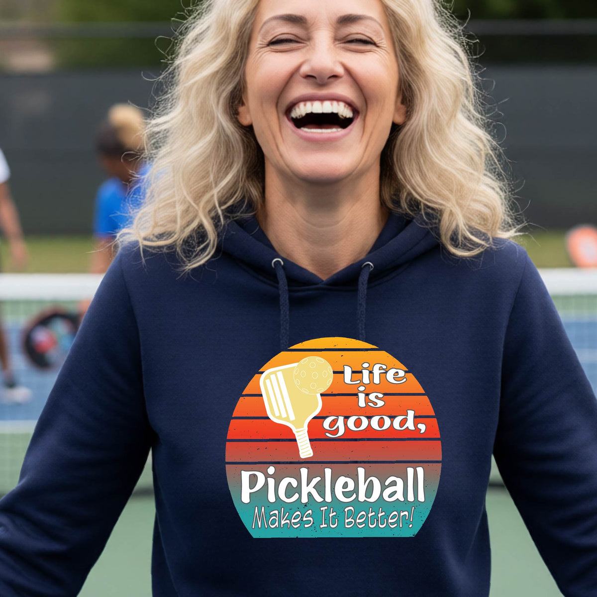 A woman wearing a navy blue hoodie pullover with a retro multi-colored circle with a pickleball paddle and ball, with white text that says 'LIFE IS GOOD PICKLEBALL MAKES IT BETTER', standing in front of a pickleball court.
