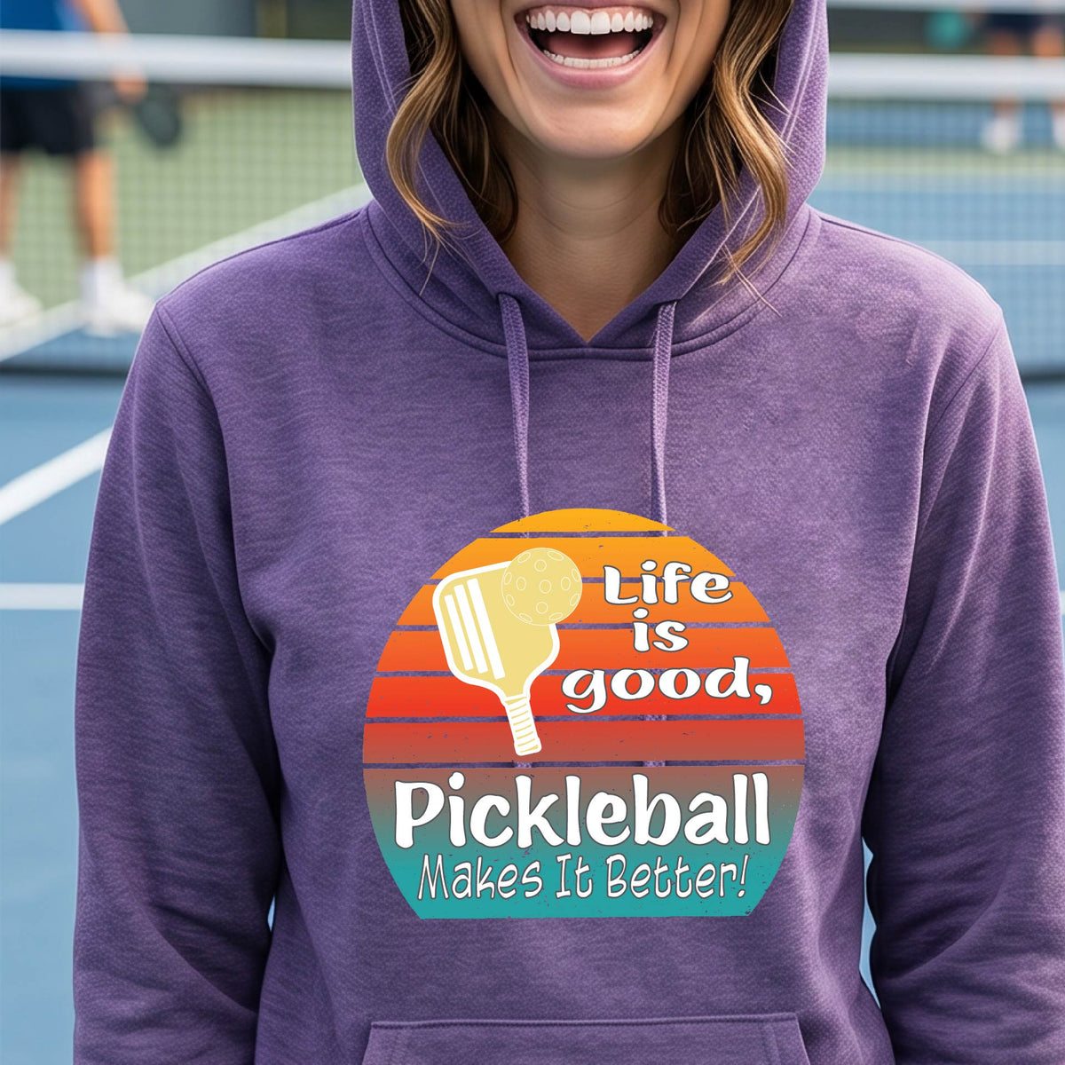 A woman wearing a purple hoodie pullover with a retro multi-colored circle with a pickleball paddle and ball, with white text that says 'LIFE IS GOOD PICKLEBALL MAKES IT BETTER', standing in front of a pickleball court.