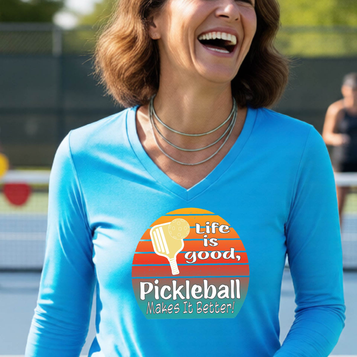 A woman wearing a bright blue long-sleeve t-shirt with a retro multi-colored circle with a pickleball paddle and ball, with white text that says 'LIFE IS GOOD PICKLEBALL MAKES IT BETTER', standing in front of a pickleball court.