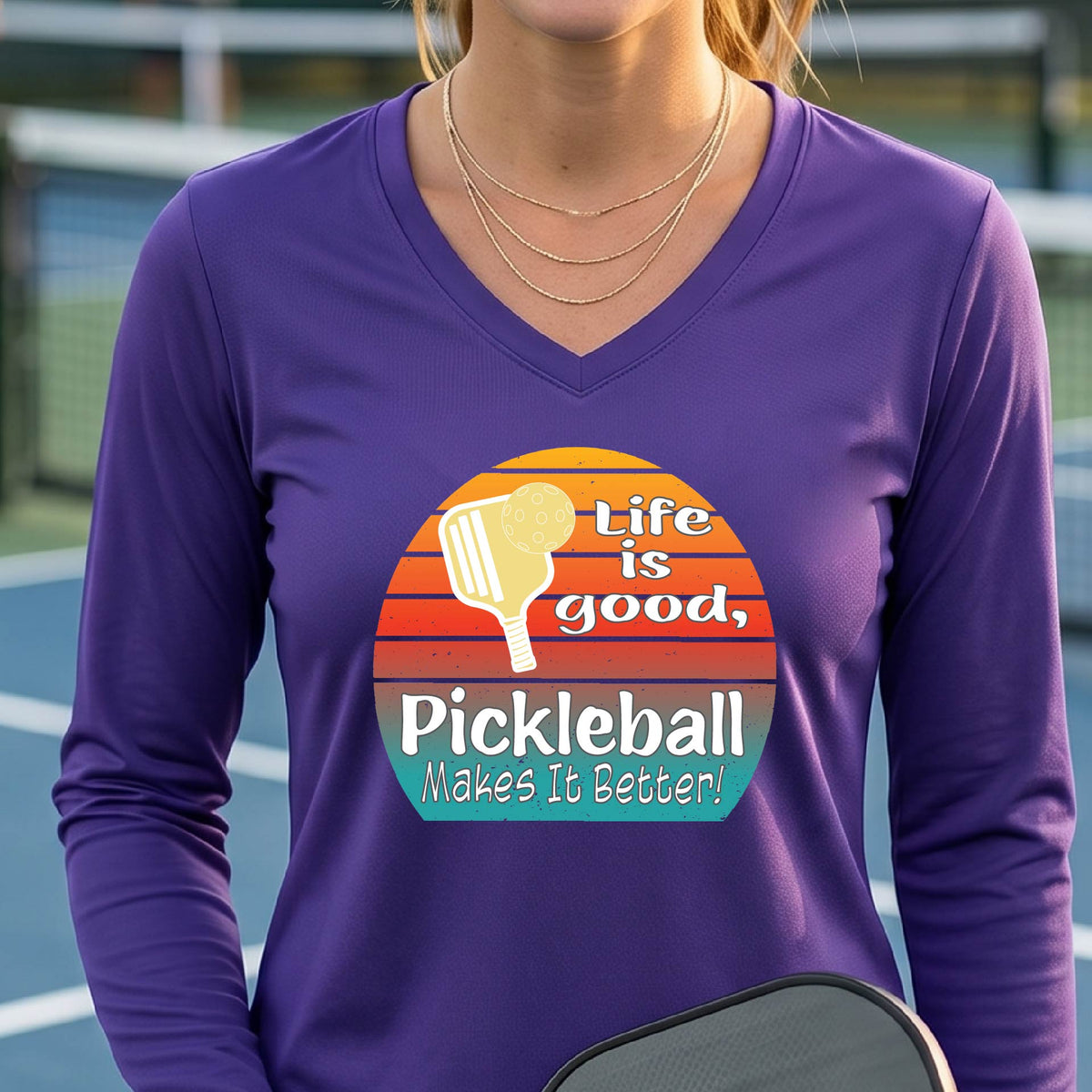 A woman wearing a purple long-sleeve t-shirt with a retro multi-colored circle with a pickleball paddle and ball, with white text that says 'LIFE IS GOOD PICKLEBALL MAKES IT BETTER', standing in front of a pickleball court.
