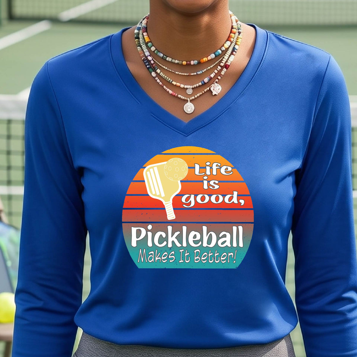 A woman wearing royal blue long-sleeve t-shirt with a retro multi-colored circle with a pickleball paddle and ball, with white text that says 'LIFE IS GOOD PICKLEBALL MAKES IT BETTER', standing in front of a pickleball court.