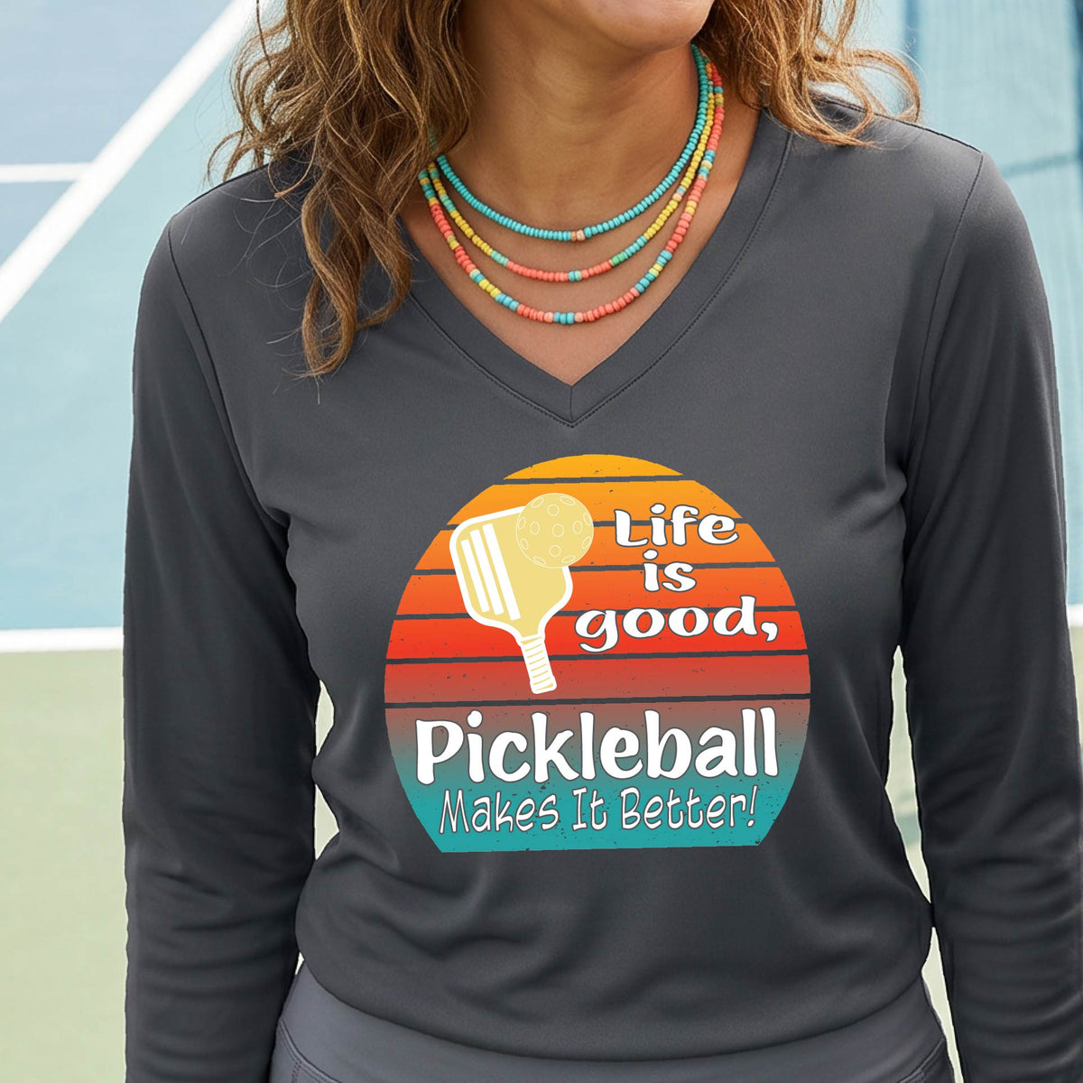 A woman wearing a dark gray long-sleeve t-shirt with a retro multi-colored circle with a pickleball paddle and ball, with white text that says 'LIFE IS GOOD PICKLEBALL MAKES IT BETTER', standing in front of a pickleball court.