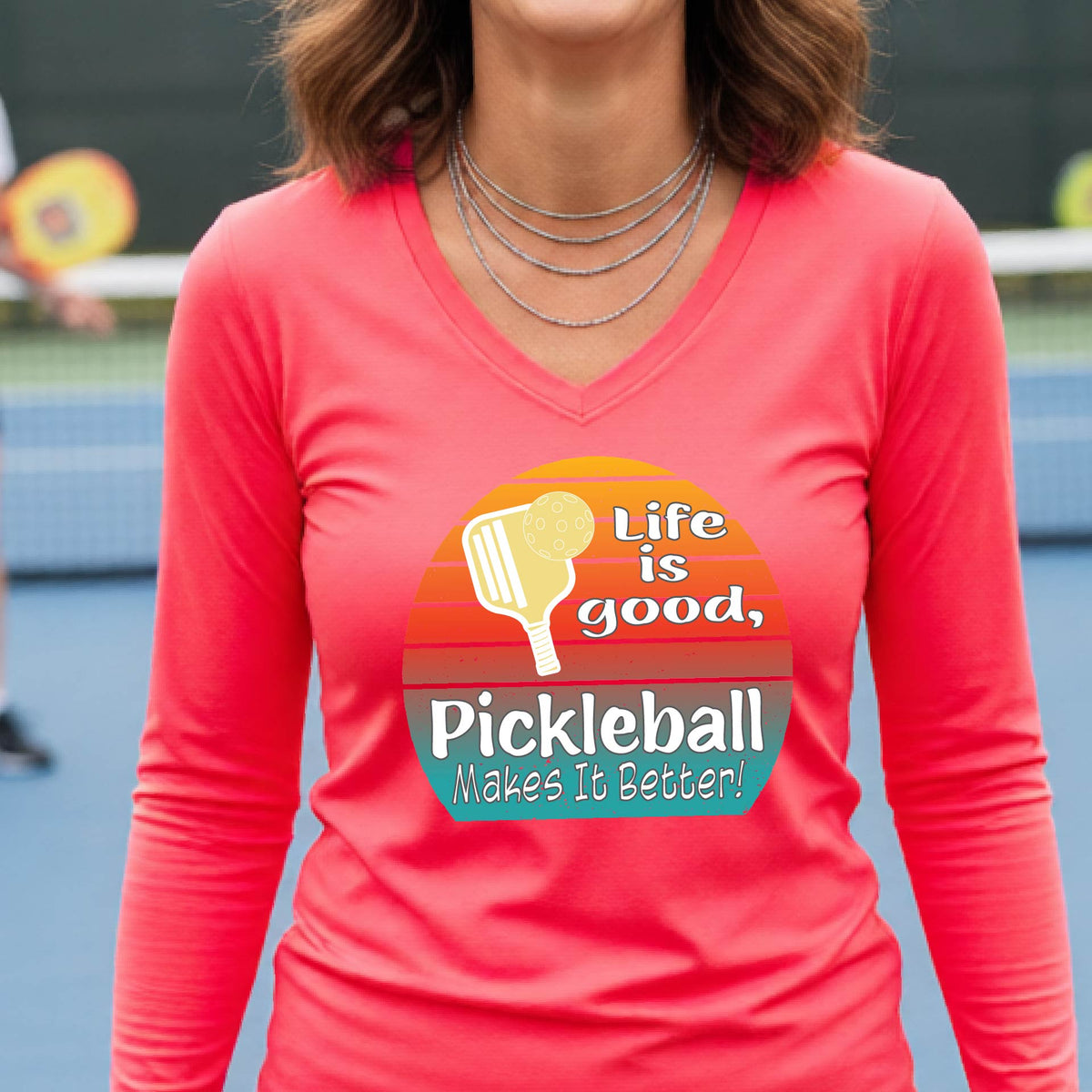 A woman wearing a coral pink long-sleeve t-shirt with a retro multi-colored circle with a pickleball paddle and ball, with white text that says 'LIFE IS GOOD PICKLEBALL MAKES IT BETTER', standing in front of a pickleball court.