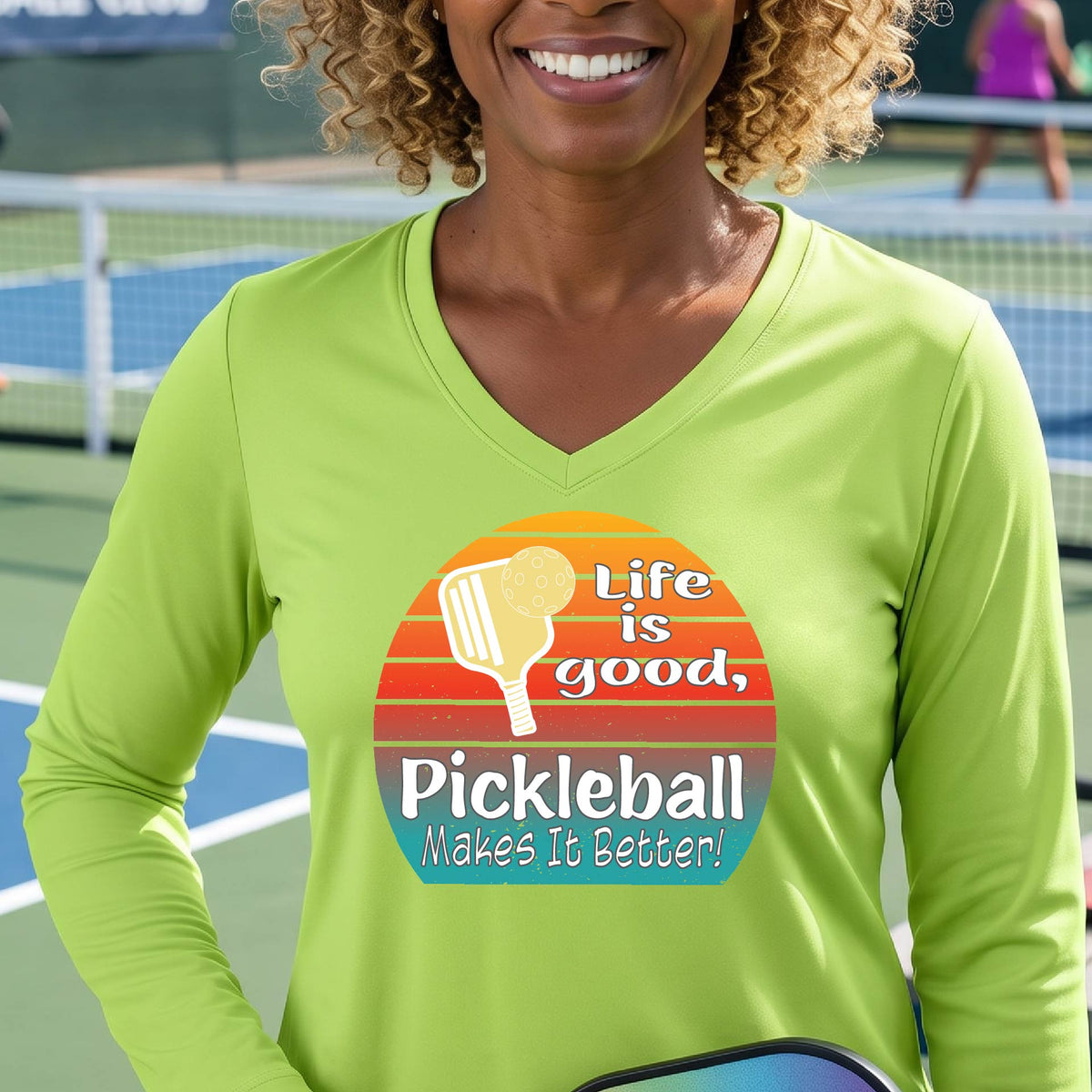 A woman wearing a lime green long-sleeve t-shirt with a retro multi-colored circle with a pickleball paddle and ball, with white text that says 'LIFE IS GOOD PICKLEBALL MAKES IT BETTER', standing in front of a pickleball court.