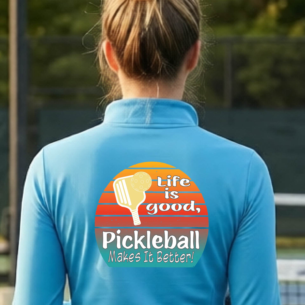 A woman wearing bright blue quarter-zip shirt with a retro multi-colored circle with a pickleball paddle and ball, with white text that says 'LIFE IS GOOD PICKLEBALL MAKES IT BETTER', standing in front of a pickleball court.