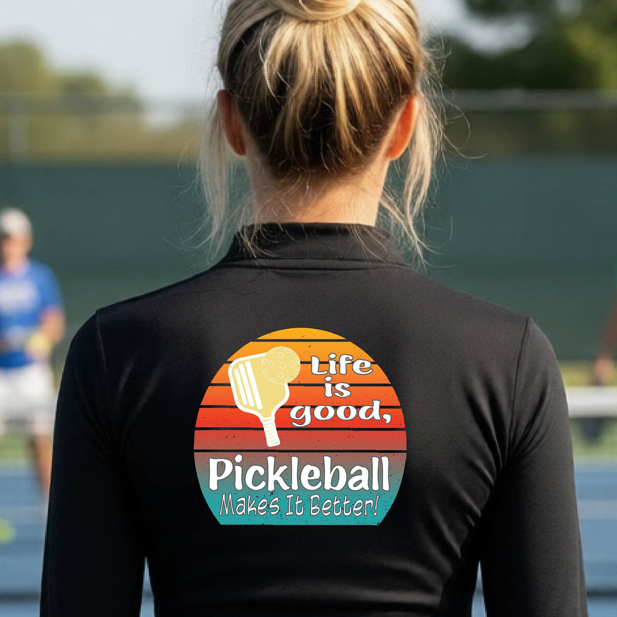 A woman wearing black quarter-zip shirt with a retro multi-colored circle with a pickleball paddle and ball, with white text that says 'LIFE IS GOOD PICKLEBALL MAKES IT BETTER', standing in front of a pickleball court.