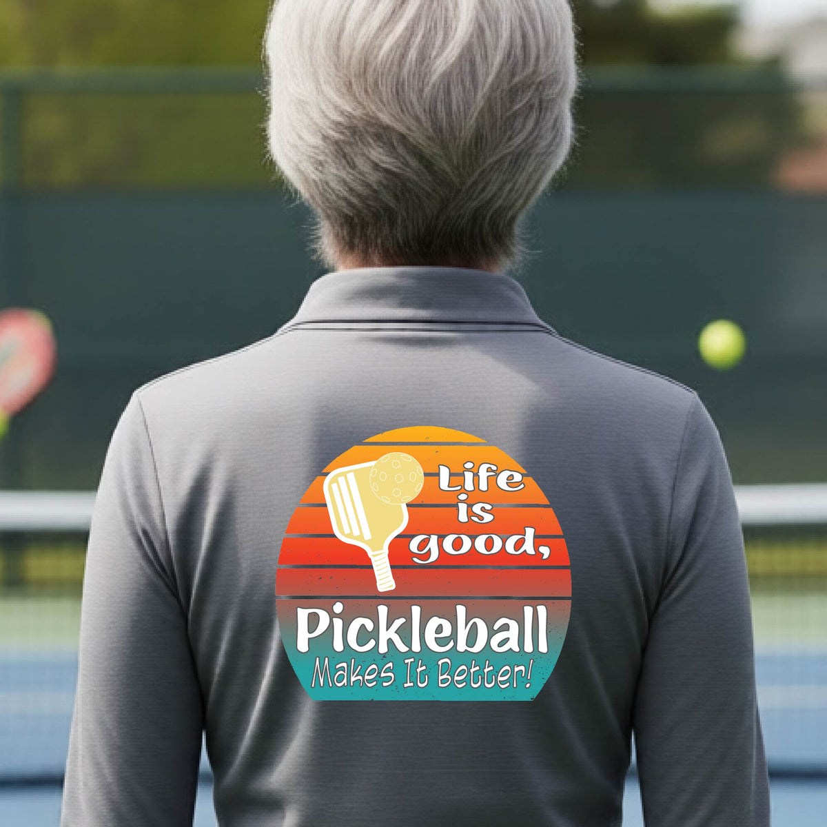 A woman wearing gray quarter-zip shirt with a retro multi-colored circle with a pickleball paddle and ball, with white text that says 'LIFE IS GOOD PICKLEBALL MAKES IT BETTER', standing in front of a pickleball court.
