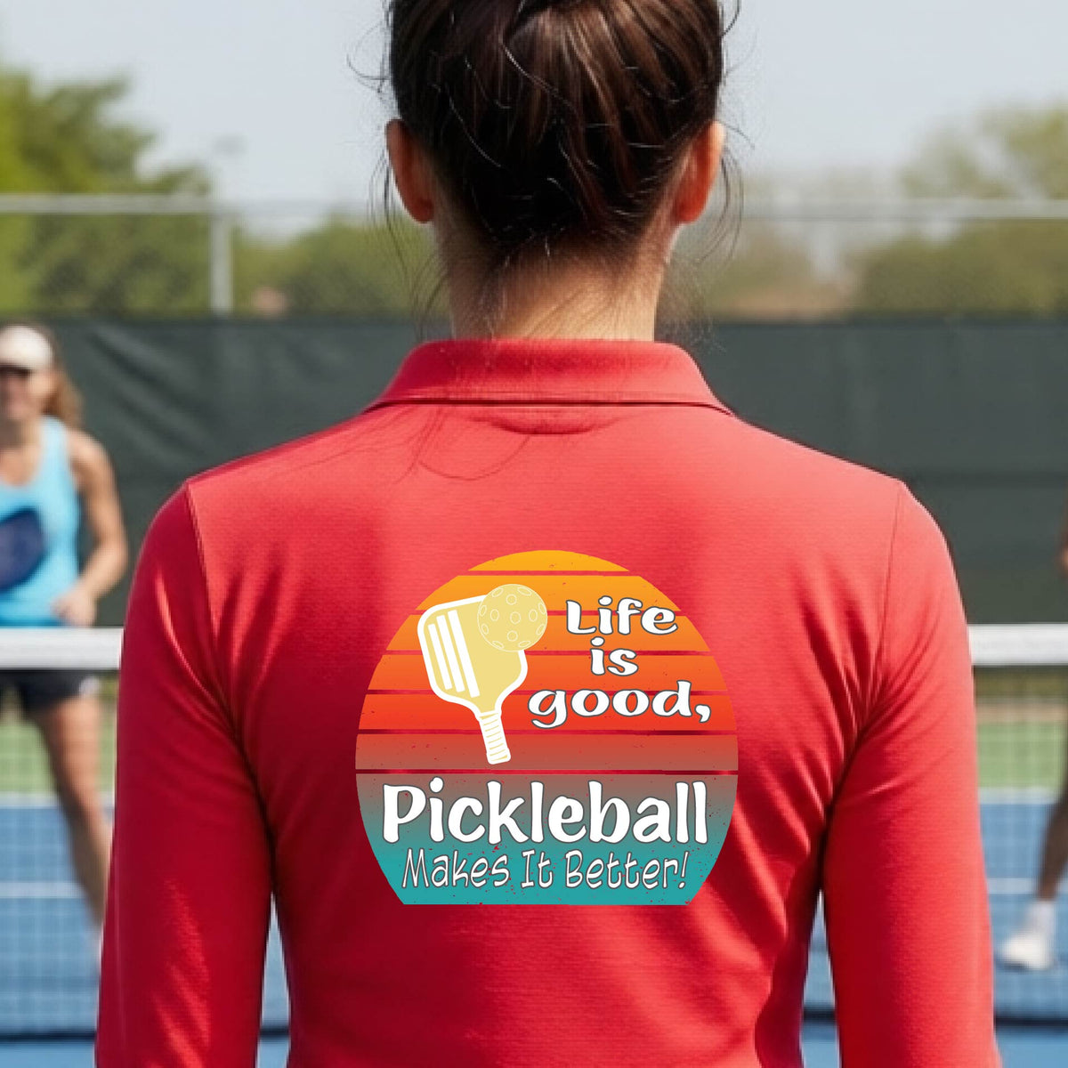 A woman wearing red quarter-zip shirt with a retro multi-colored circle with a pickleball paddle and ball, with white text that says 'LIFE IS GOOD PICKLEBALL MAKES IT BETTER', standing in front of a pickleball court.