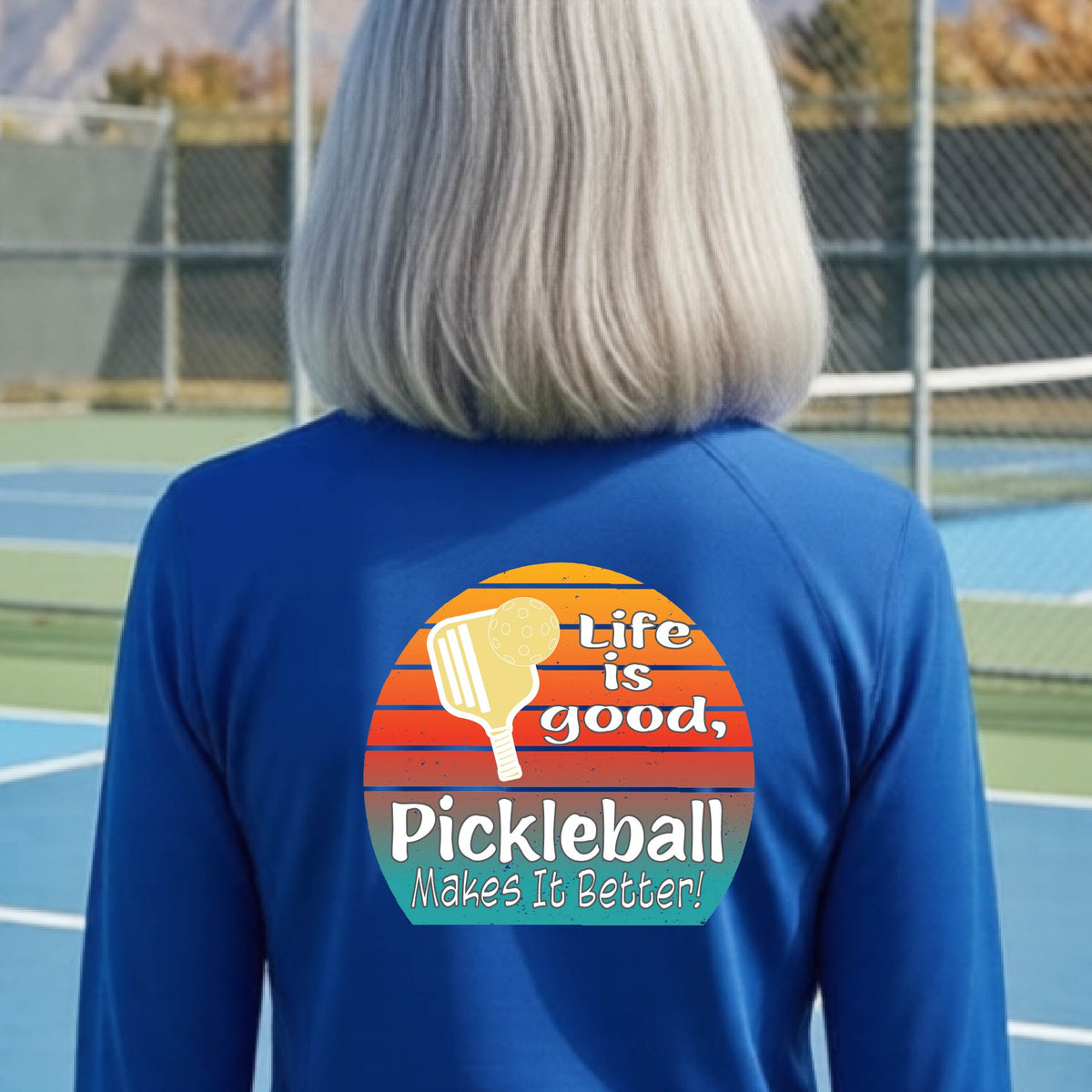 A woman wearing royal blue quarter-zip shirt with a retro multi-colored circle with a pickleball paddle and ball, with white text that says 'LIFE IS GOOD PICKLEBALL MAKES IT BETTER', standing in front of a pickleball court.