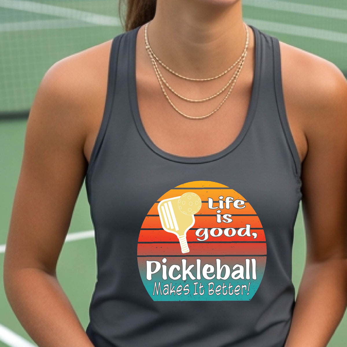 A woman wearing a dark gray racerback tank top with a retro multi-colored circle with a pickleball paddle and ball, with white text that says 'LIFE IS GOOD PICKLEBALL MAKES IT BETTER', standing in front of a pickleball court.