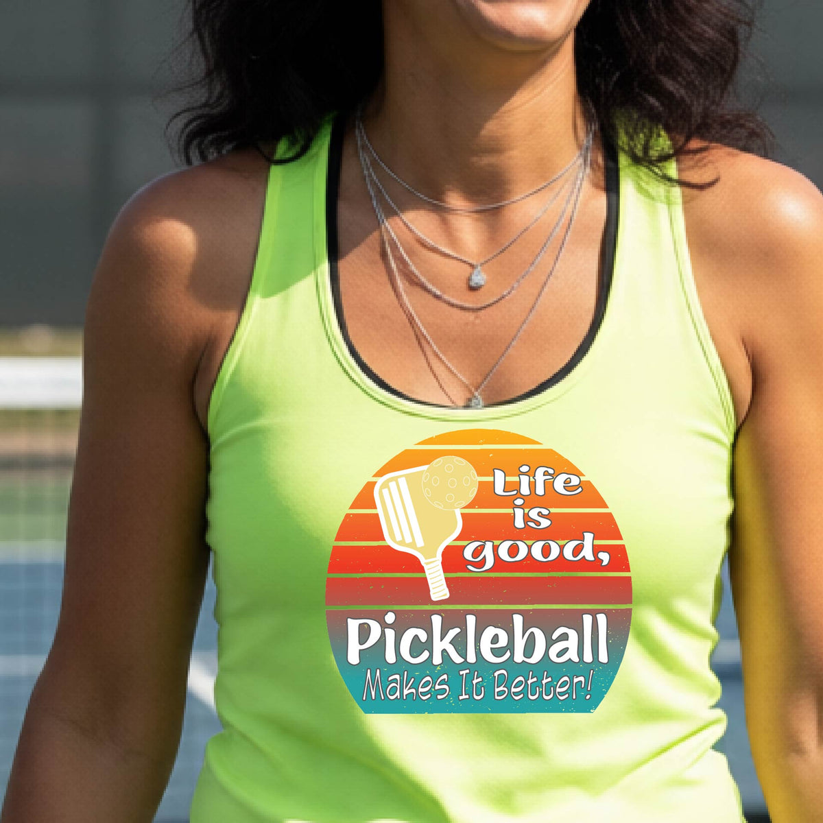 A woman wearing a lime green racerback tank top with a retro multi-colored circle with a pickleball paddle and ball, with white text that says 'LIFE IS GOOD PICKLEBALL MAKES IT BETTER', standing in front of a pickleball court.