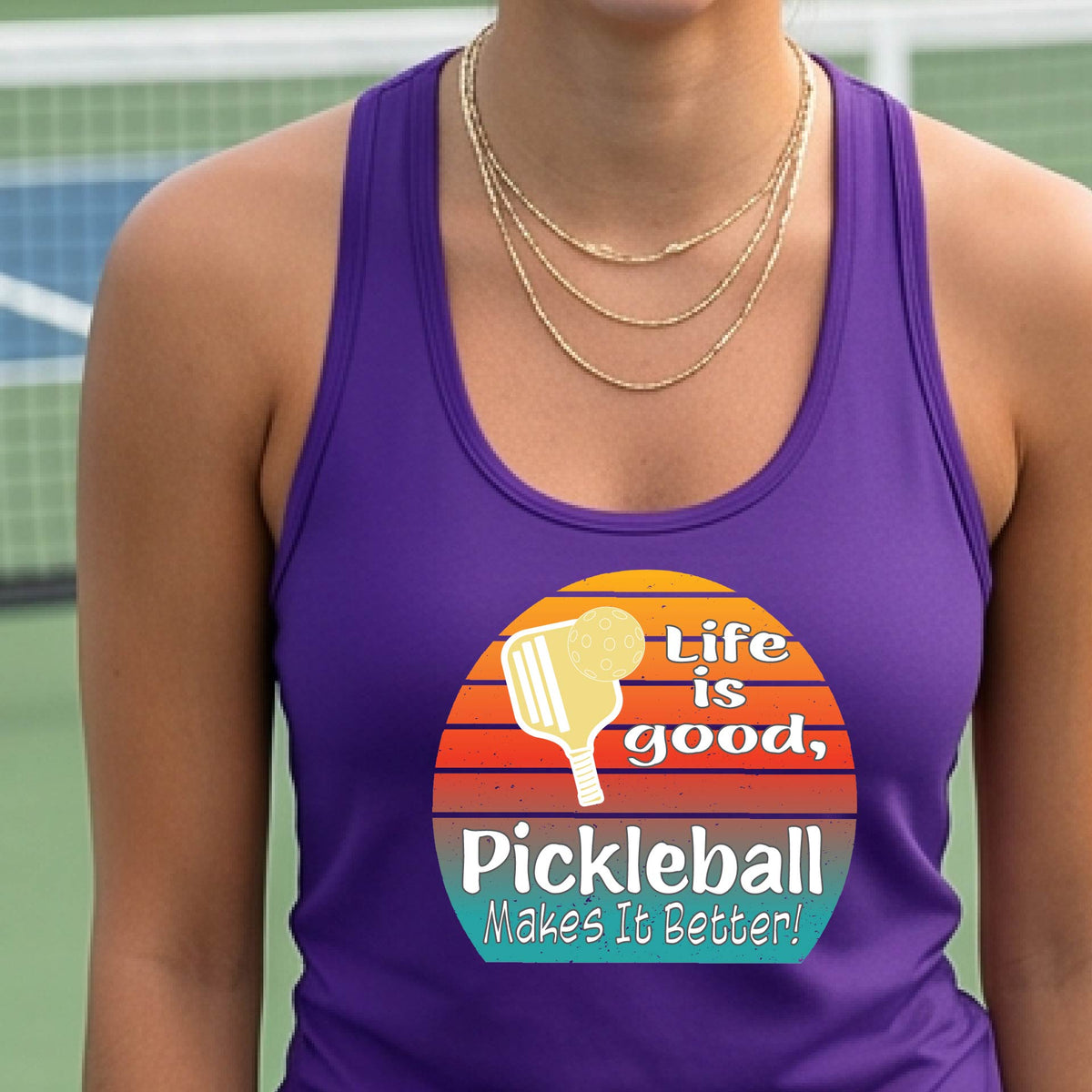 A woman wearing a purple racerback tank top with a retro multi-colored circle with a pickleball paddle and ball, with white text that says 'LIFE IS GOOD PICKLEBALL MAKES IT BETTER', standing in front of a pickleball court.
