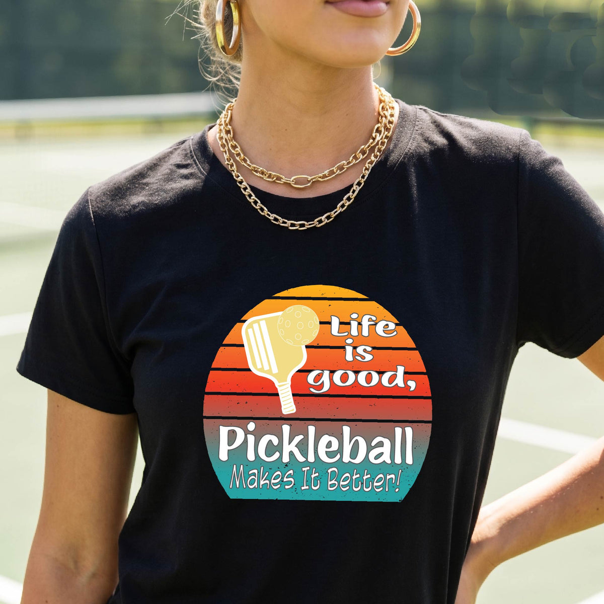 A woman wearing a black short-sleeve t-shirt with a retro multi-colored circle with a pickleball paddle and ball, with white text that says 'LIFE IS GOOD PICKLEBALL MAKES IT BETTER', standing in front of a pickleball court. 