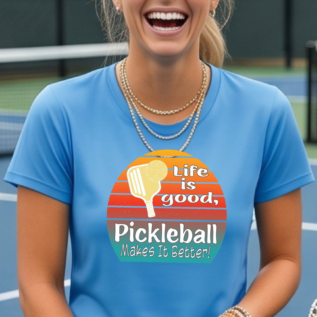 A woman wearing a light blue short-sleeve t-shirt with a retro multi-colored circle with a pickleball paddle and ball, with white text that says 'LIFE IS GOOD PICKLEBALL MAKES IT BETTER', standing in front of a pickleball court. 