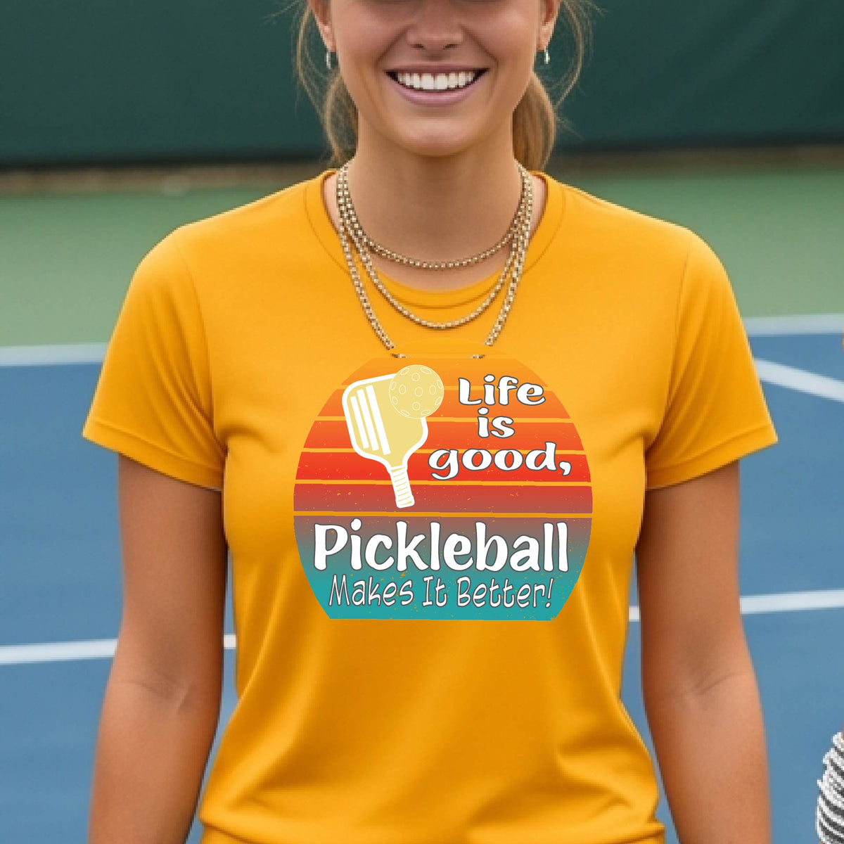 A woman wearing a mustard yellow gold short-sleeve t-shirt with a retro multi-colored circle with a pickleball paddle and ball, with white text that says 'LIFE IS GOOD PICKLEBALL MAKES IT BETTER', standing in front of a pickleball court. 