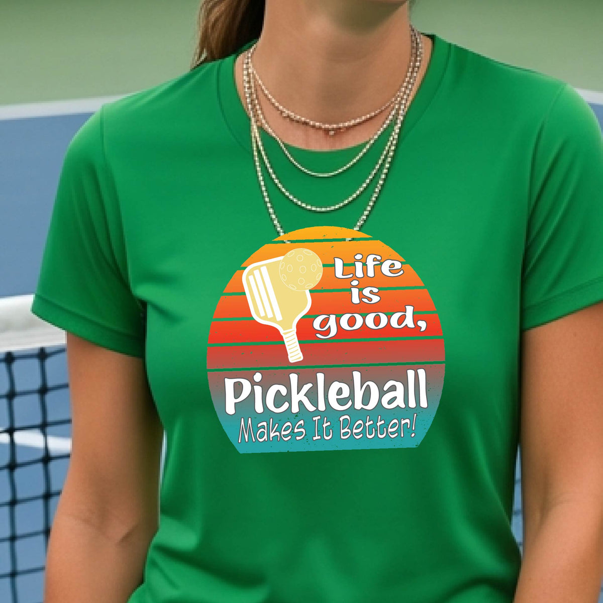 A woman wearing a green short-sleeve t-shirt with a retro multi-colored circle with a pickleball paddle and ball, with white text that says 'LIFE IS GOOD PICKLEBALL MAKES IT BETTER', standing in front of a pickleball court. 