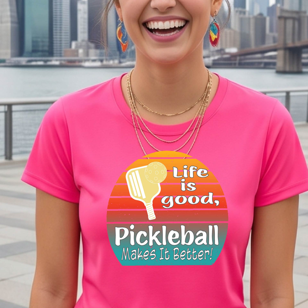 A woman wearing a pink short-sleeve t-shirt with a retro multi-colored circle with a pickleball paddle and ball, with white text that says 'LIFE IS GOOD PICKLEBALL MAKES IT BETTER', standing in front of a pickleball court. 
