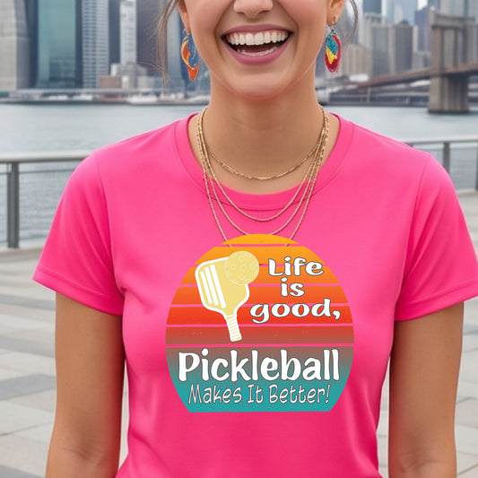 A woman wearing a pink short-sleeve t-shirt with a retro multi-colored circle with a pickleball paddle and ball, with white text that says 'LIFE IS GOOD PICKLEBALL MAKES IT BETTER', standing in front of a pickleball court. 