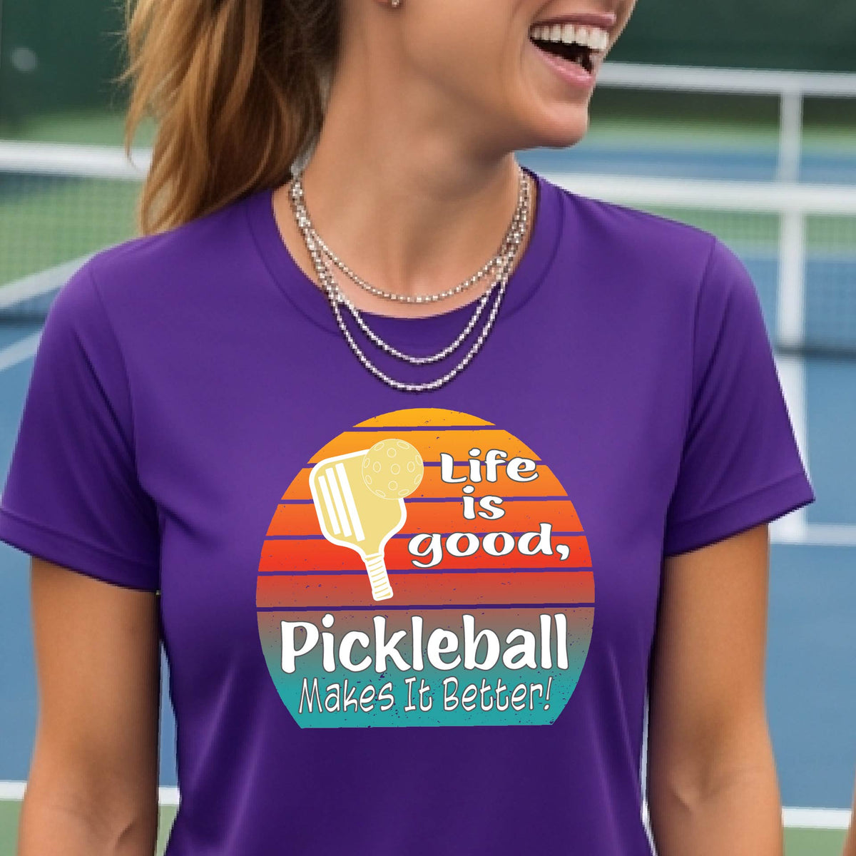 A woman wearing a purple short-sleeve t-shirt with a retro multi-colored circle with a pickleball paddle and ball, with white text that says 'LIFE IS GOOD PICKLEBALL MAKES IT BETTER', standing in front of a pickleball court. 