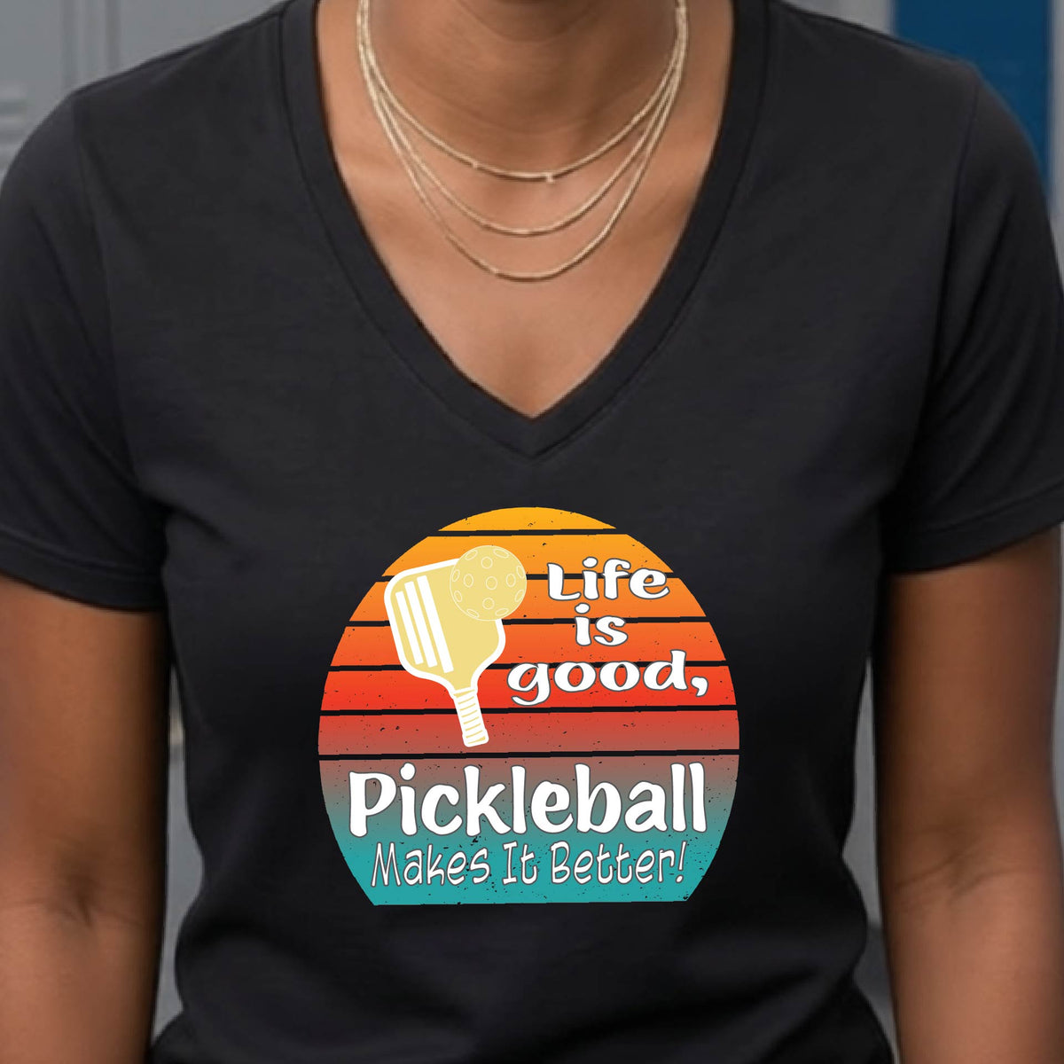 A person wearing a black V-neck t-shirt with a retro-style pickleball graphic on the chest. The graphic features a yellow pickleball paddle, a red, orange, and yellow sunset gradient, and the text 'Life is good, Pickleball Makes It Better!'.
