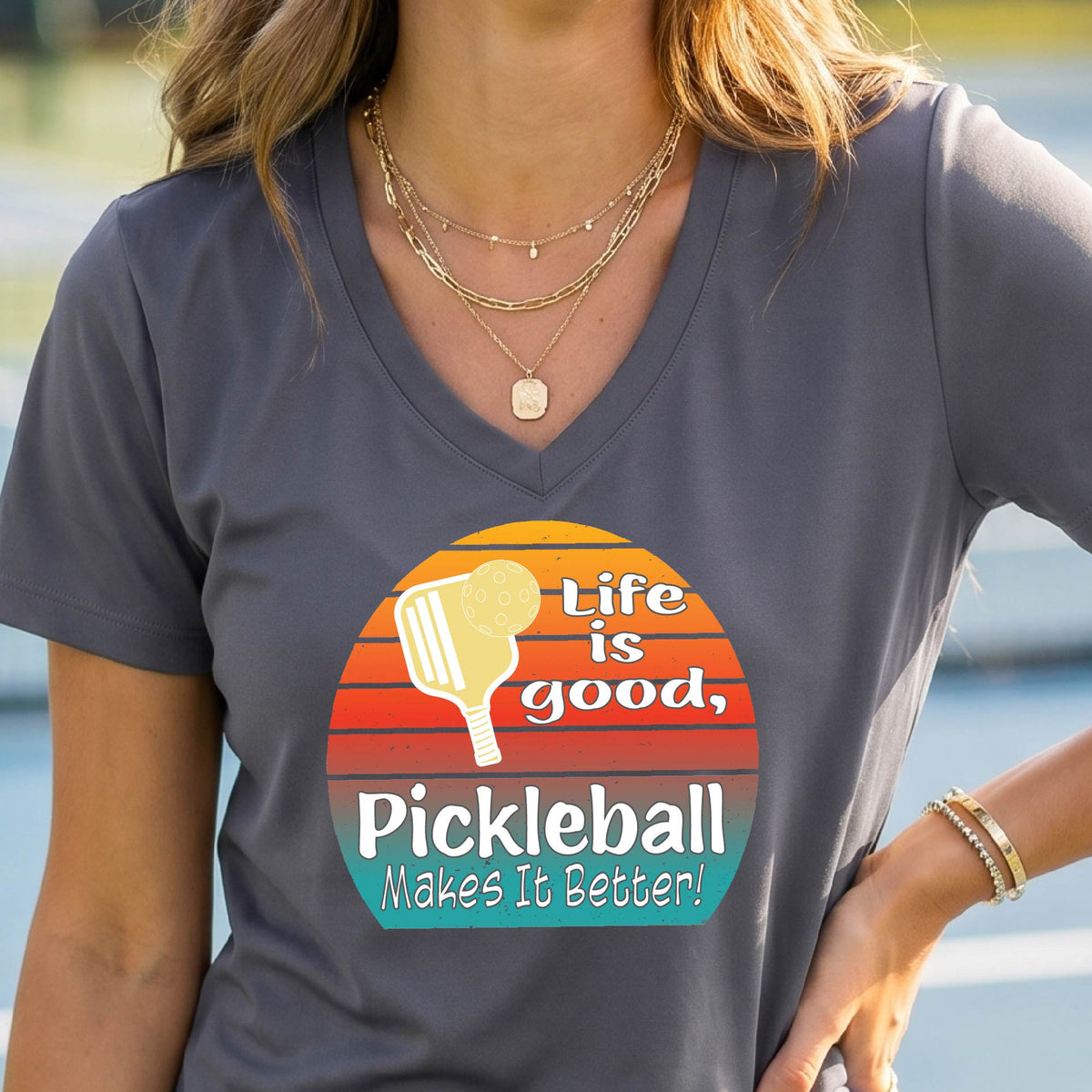 A person wearing a dark gray V-neck t-shirt with a retro-style pickleball graphic on the chest. The graphic features a yellow pickleball paddle, a red, orange, and yellow sunset gradient, and the text 'Life is good, Pickleball Makes It Better'.