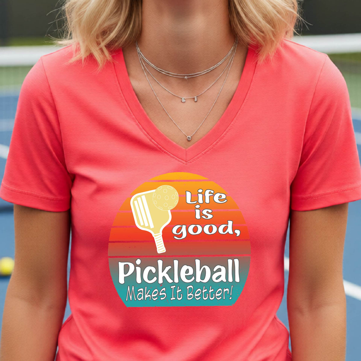 A person wearing a coral pink V-neck t-shirt with a retro-style pickleball graphic on the chest. The graphic features a yellow pickleball paddle, a red, orange, and yellow sunset gradient, and the text 'Life is good, Pickleball Makes It Better!'.