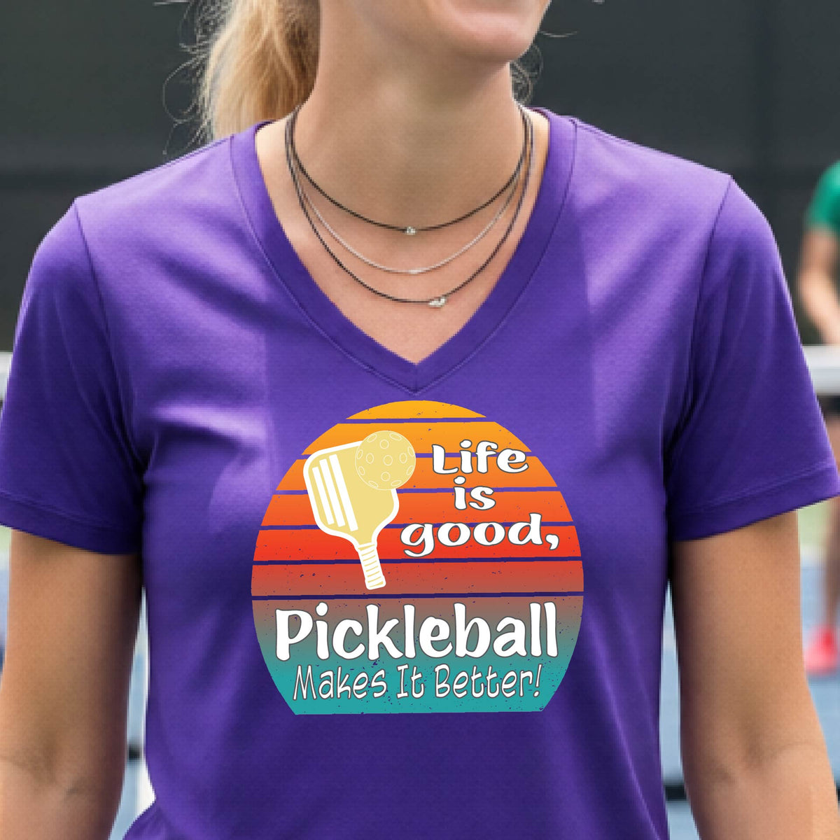 A person wearing a purple V-neck t-shirt with a retro-style pickleball graphic on the chest. The graphic features a yellow pickleball paddle, a red, orange, and yellow sunset gradient, and the text 'Life is good, Pickleball Makes It Better!'.