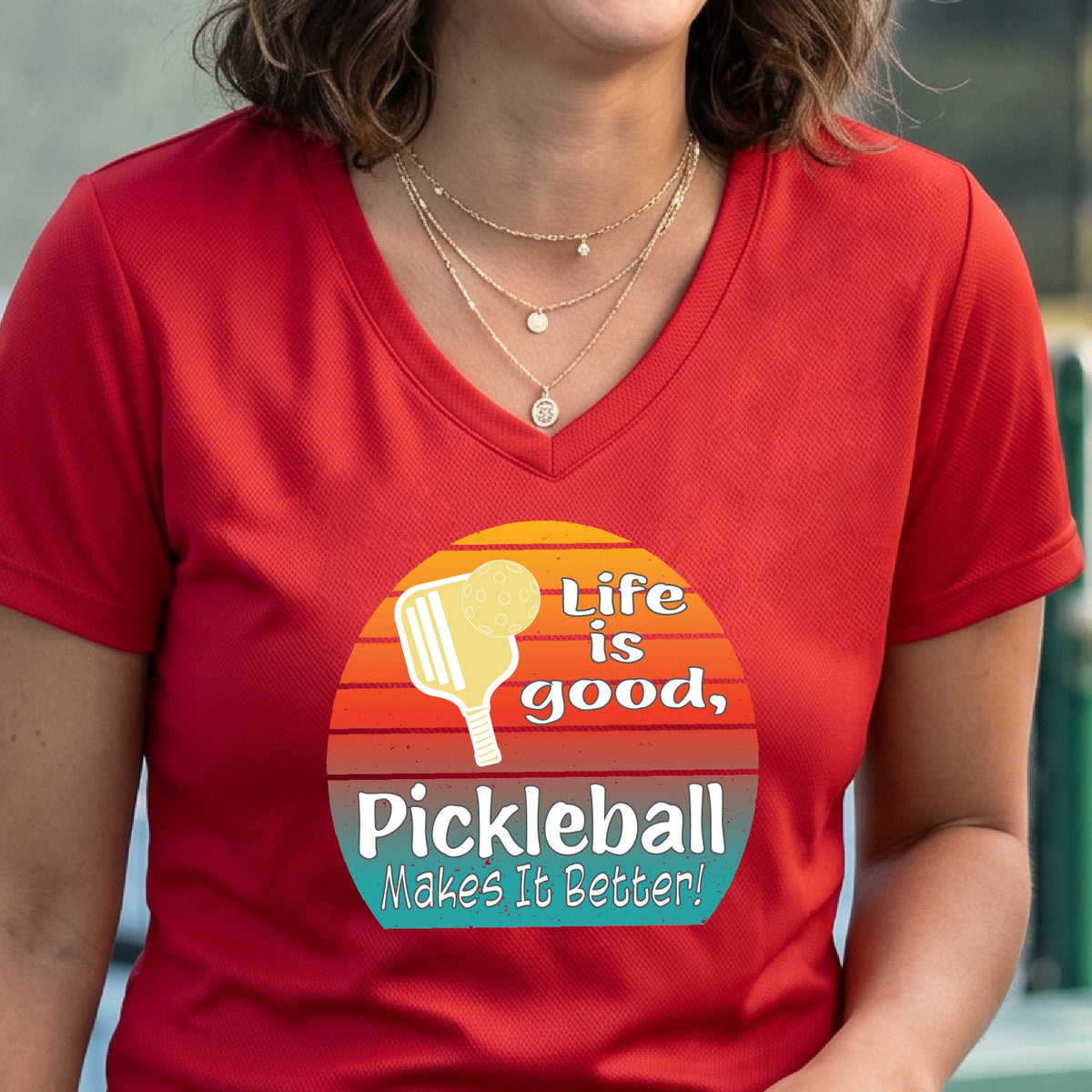 A person wearing a red V-neck t-shirt with a retro-style pickleball graphic on the chest. The graphic features a yellow pickleball paddle, a red, orange, and yellow sunset gradient, and the text 'Life is good, Pickleball Makes It Better!'.